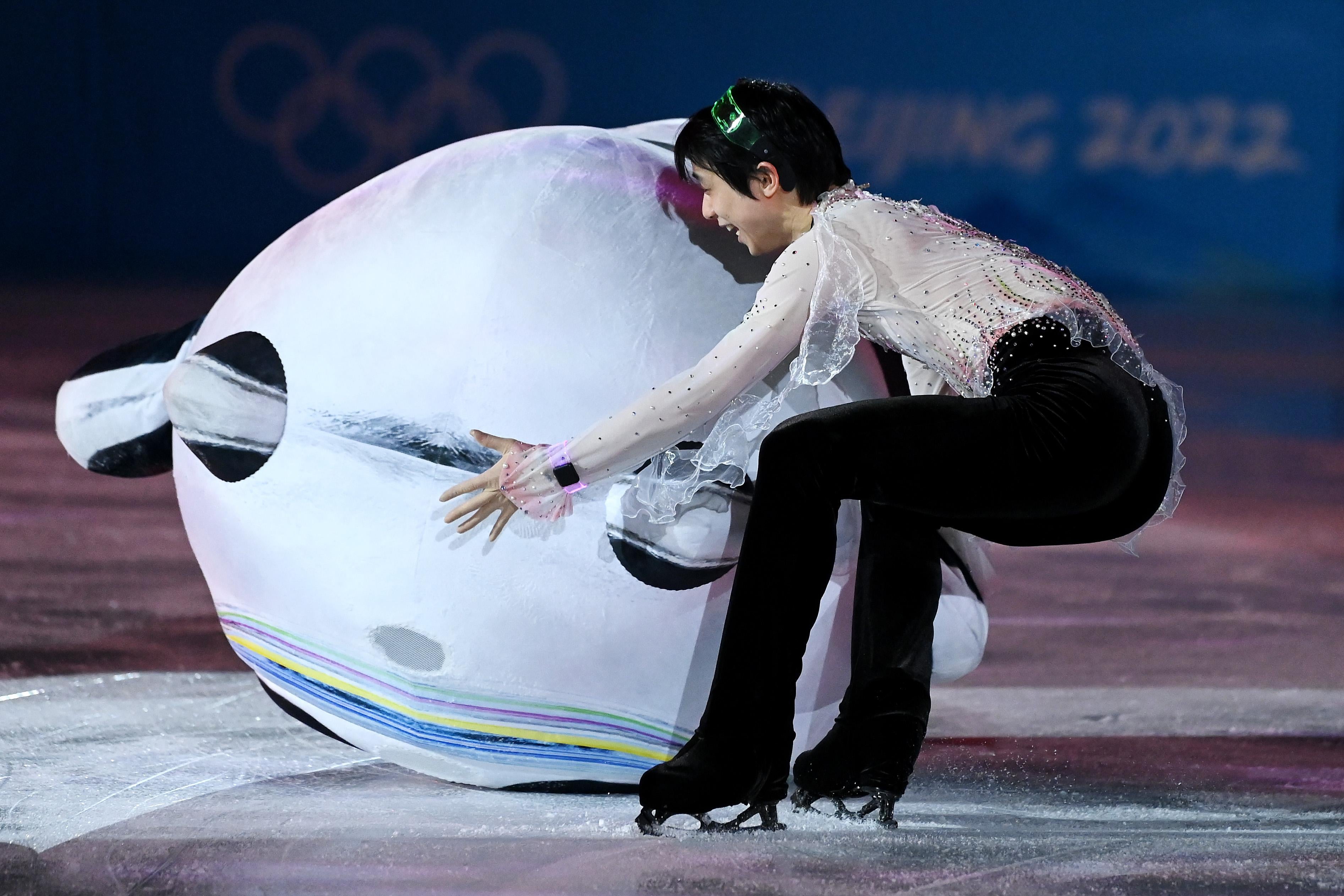 Bing Dwen Dwen face down on the ice, Hanyu crouching with his arms around him and trying to help him up.