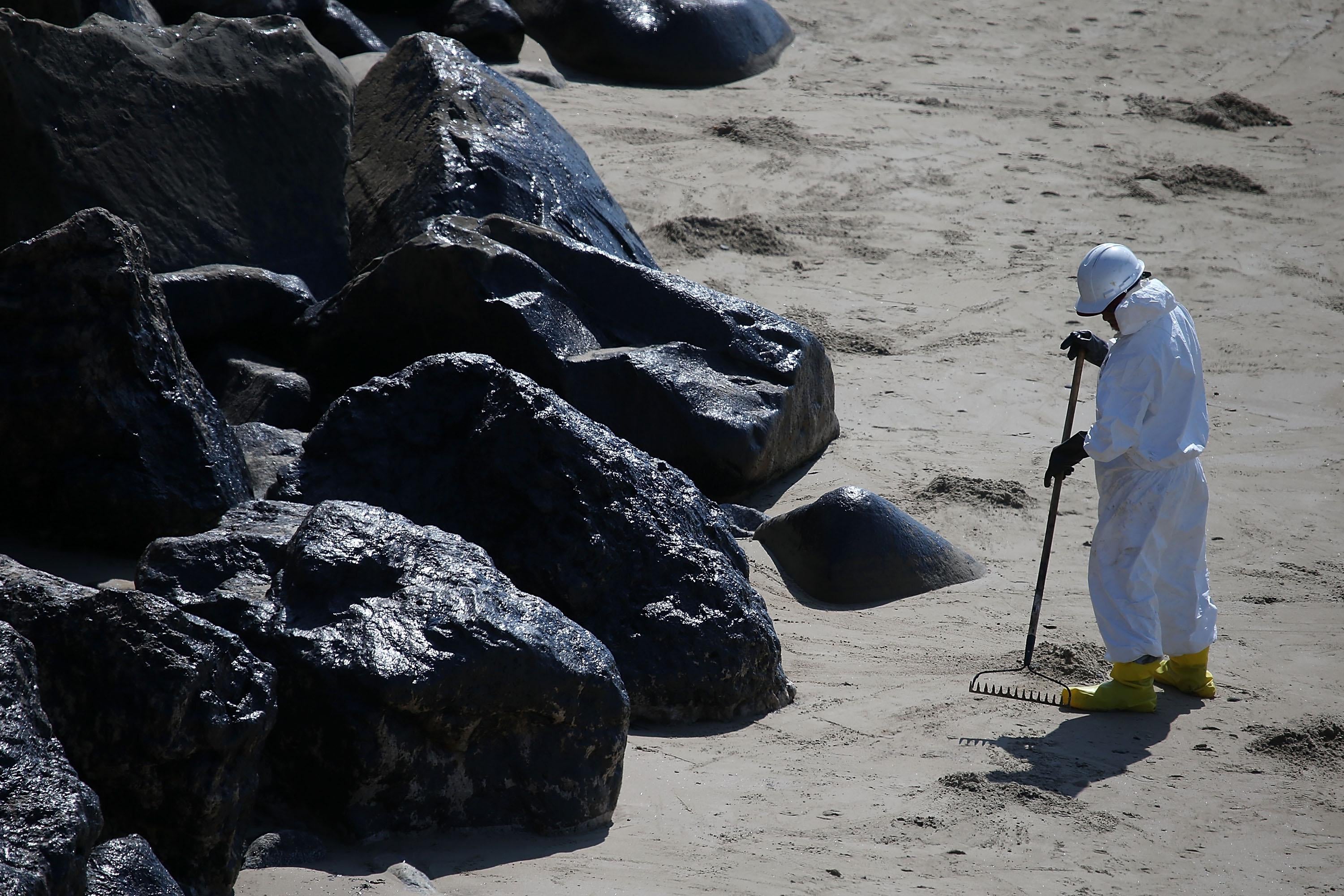 A worker in hazmat gear rakes sand next to oil-covered rocks on a beach.