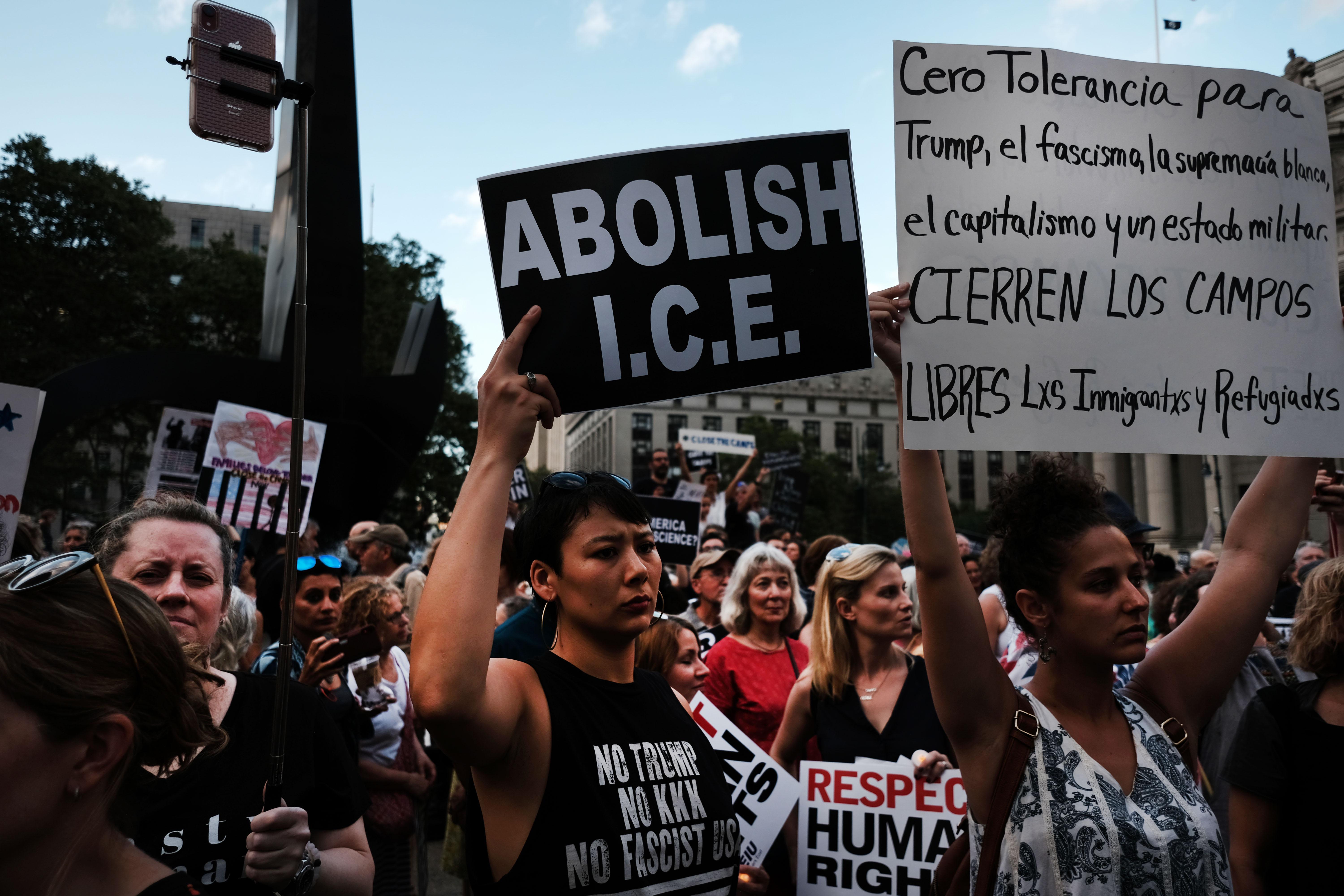 Protesters hold up signs during the daytime. The sign in the middle reads, "ABOLISH I.C.E." A woman holds up a sign in Spanish to the right.