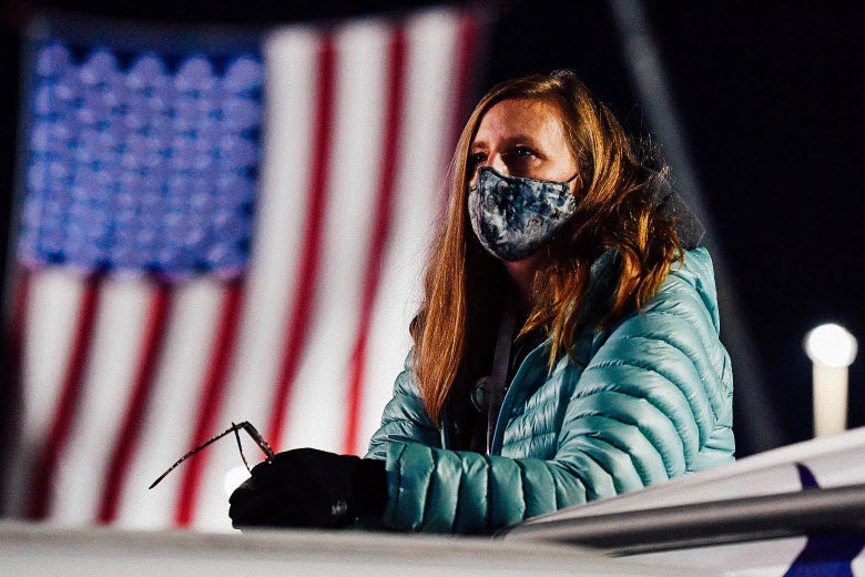 A woman wearing a mask and a puffer jacket leans on a railing outside, looking up anxiously