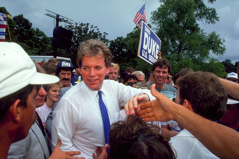David duke in shirt and tie surrounded by people shaking hands. One man wears a Duke hat; another waves a Duke for Senate sign.