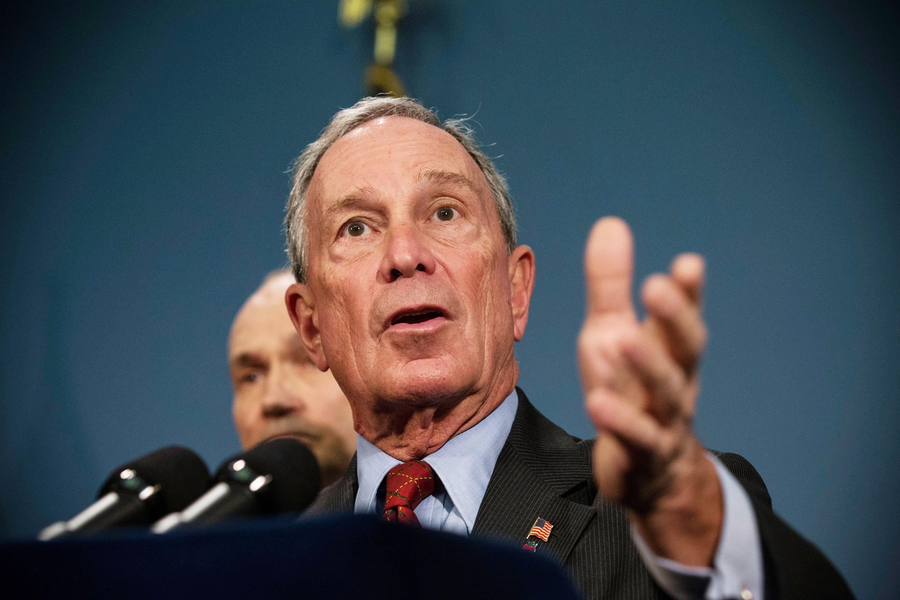 Michael Bloomberg speaks into a microphone and raises his hand.
