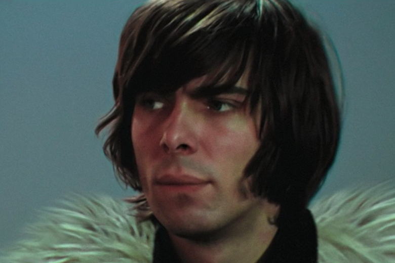 A man with brown chin-length hair wearing a top with white feathers on it