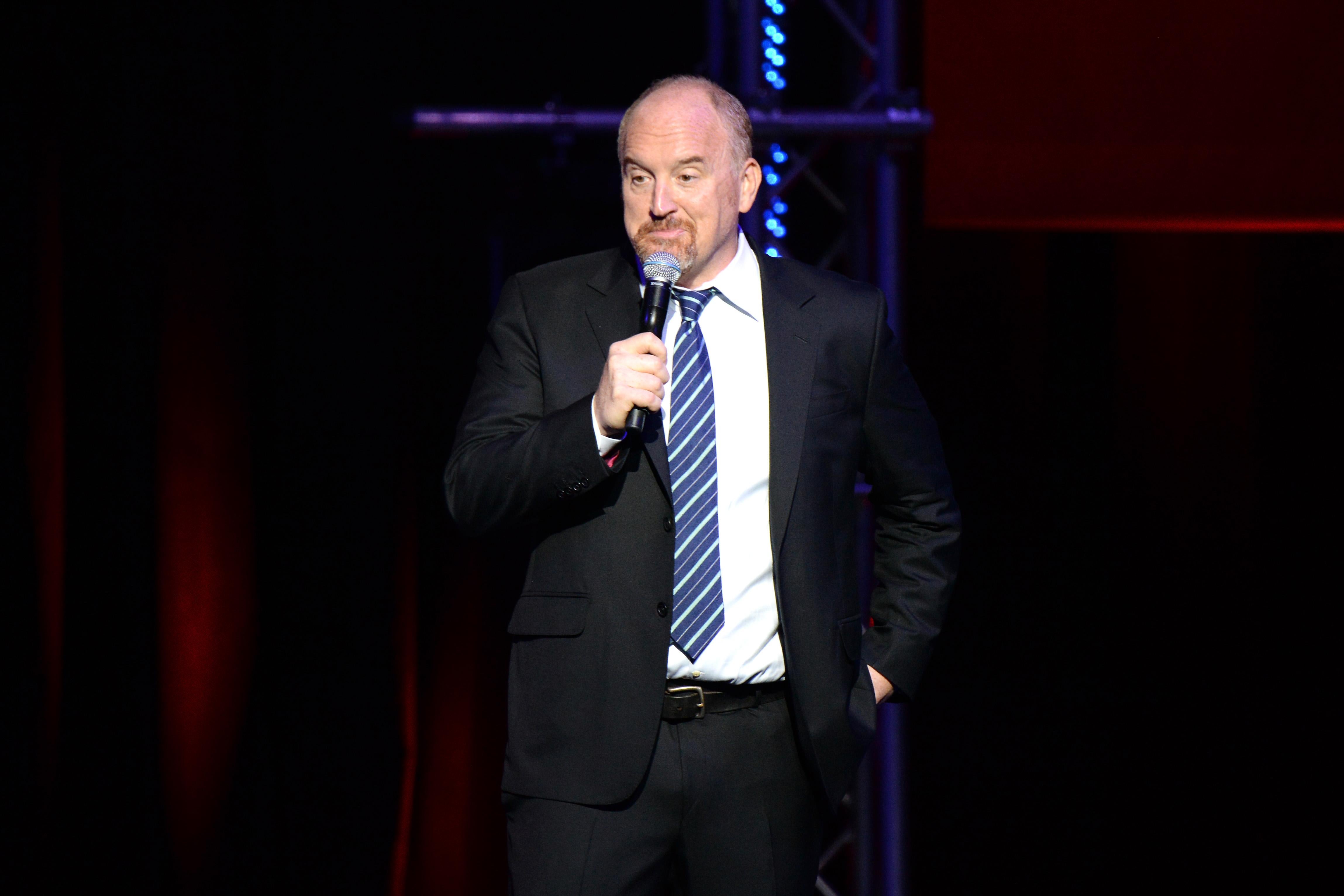 Louis C.K. wearing a suit with a blue tie, with a microphone in his right hand