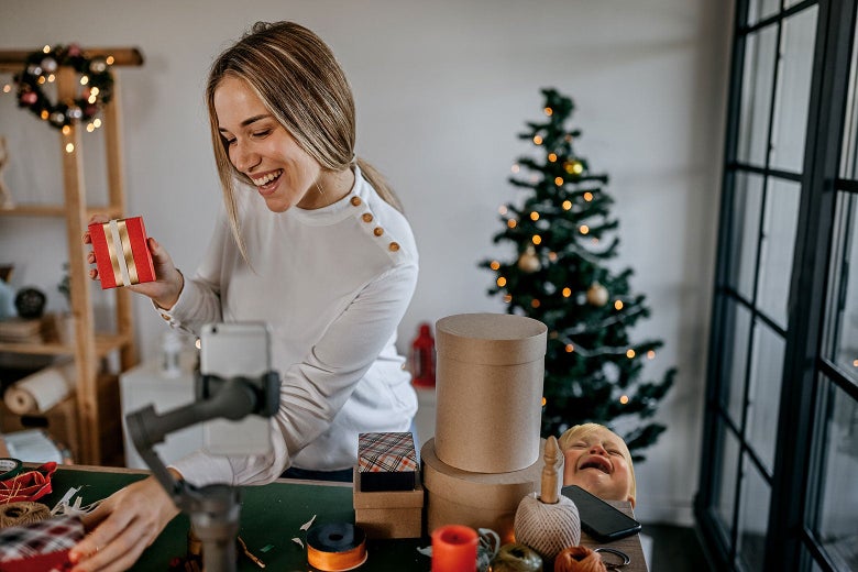 A thirtysomething woman smiles and holds up a gift box in front of a mounted smartphone recording the scene full of Christmas decorations. A baby wails beside the woman.