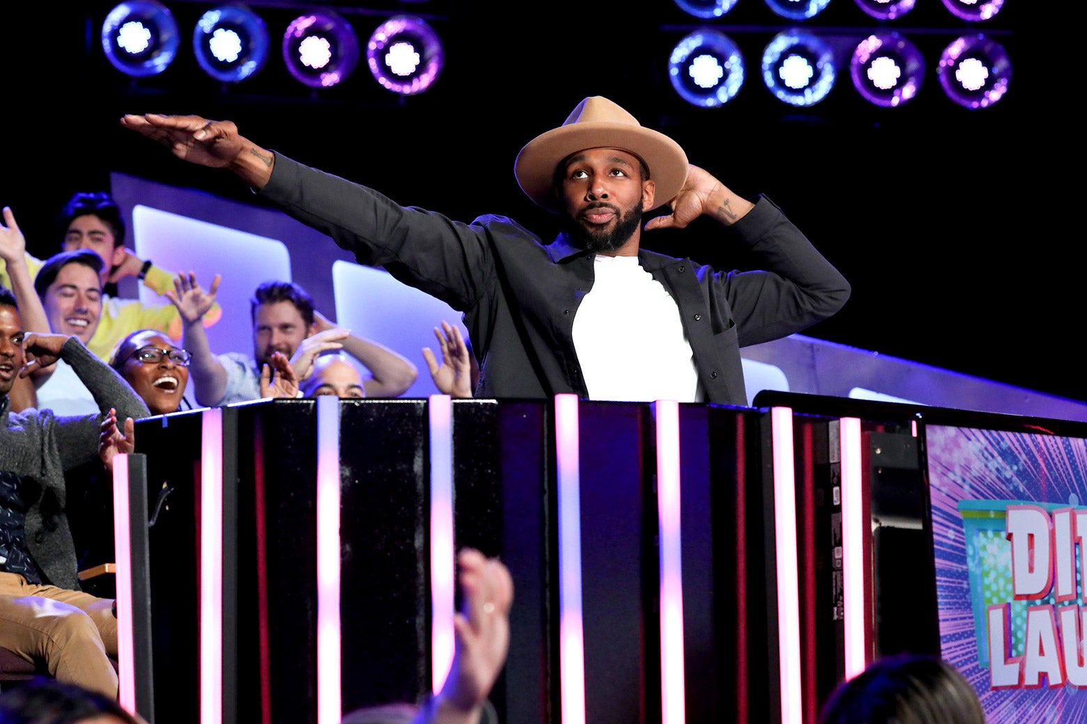 A man in a hat points to the side while happy fans smile behind him. 