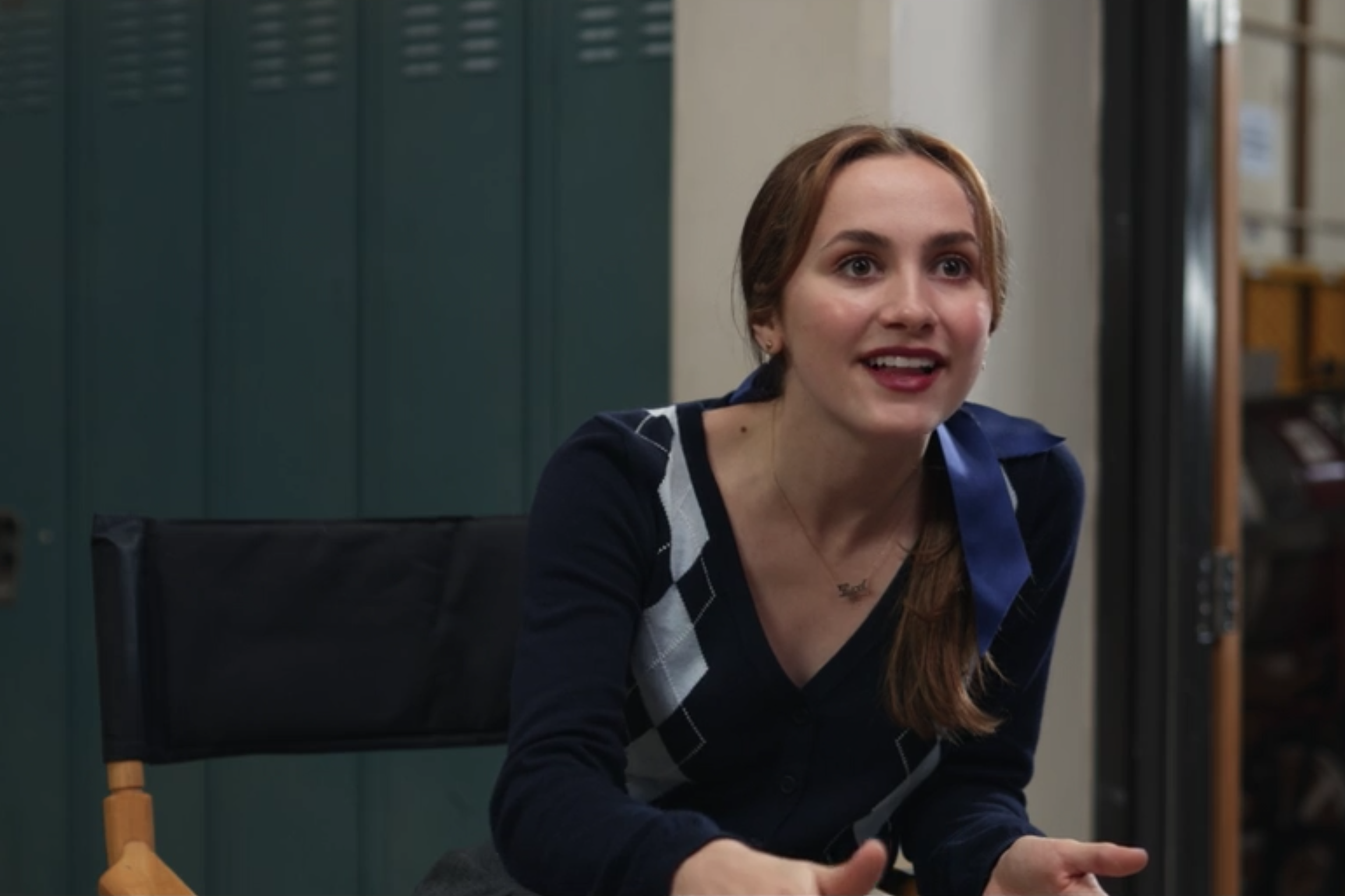 A girl sits in a director's chair wearing an argyle sweater, smiling. 