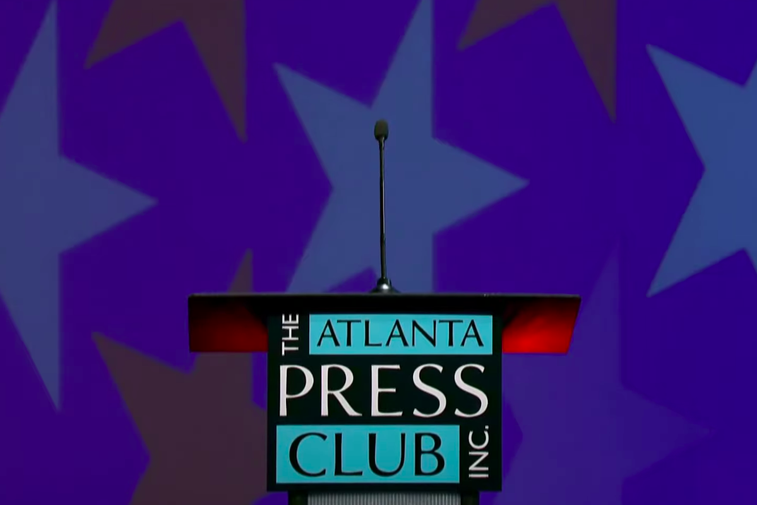 A microphone emerges atop on a podium that says the Atlanta Press Club Inc. It is in front of a blue background with large red and white stars. 