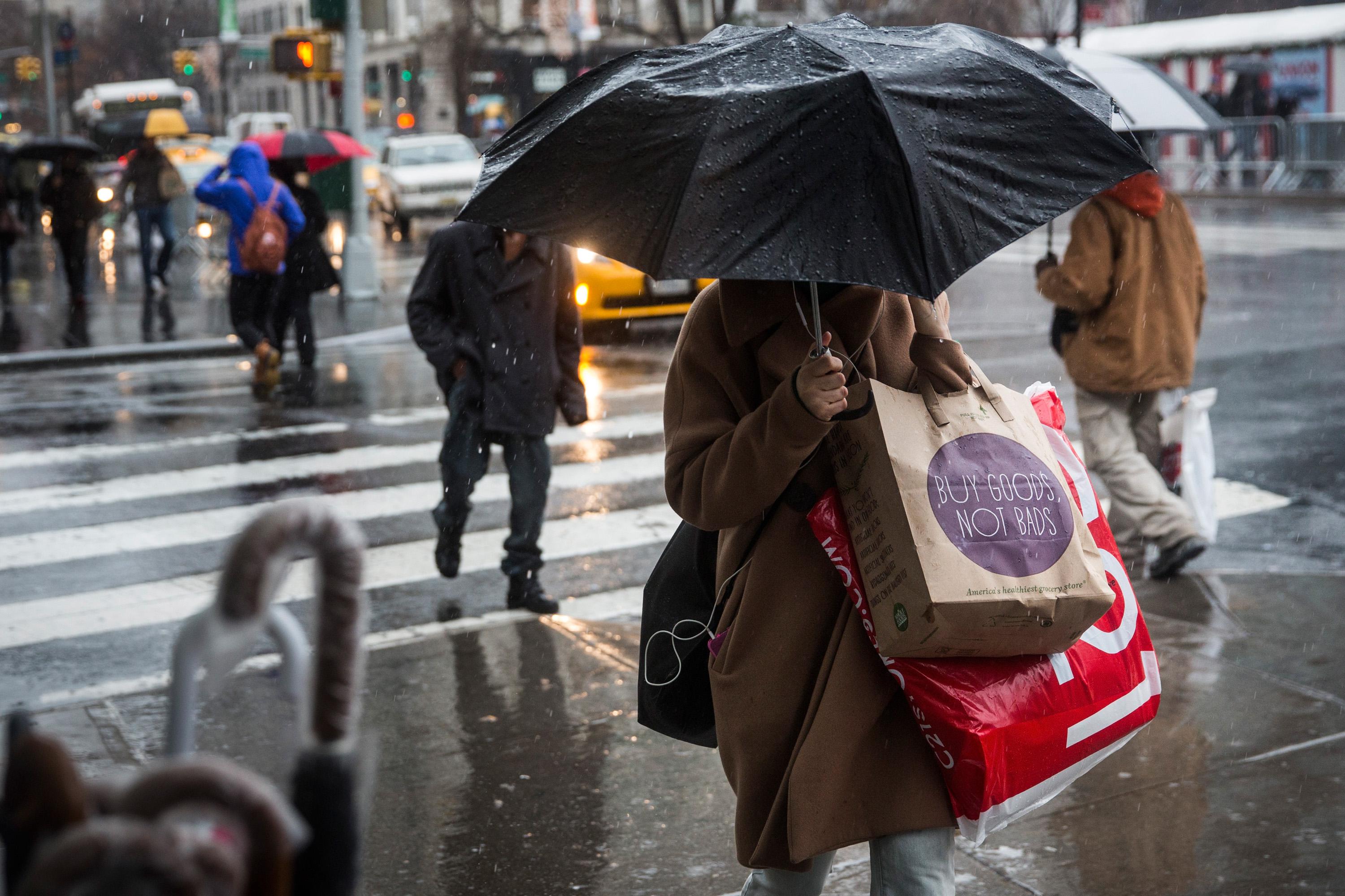 A woman crosses the street in the rain, a black umbrella covering her face. She is carrying several shopping bags.