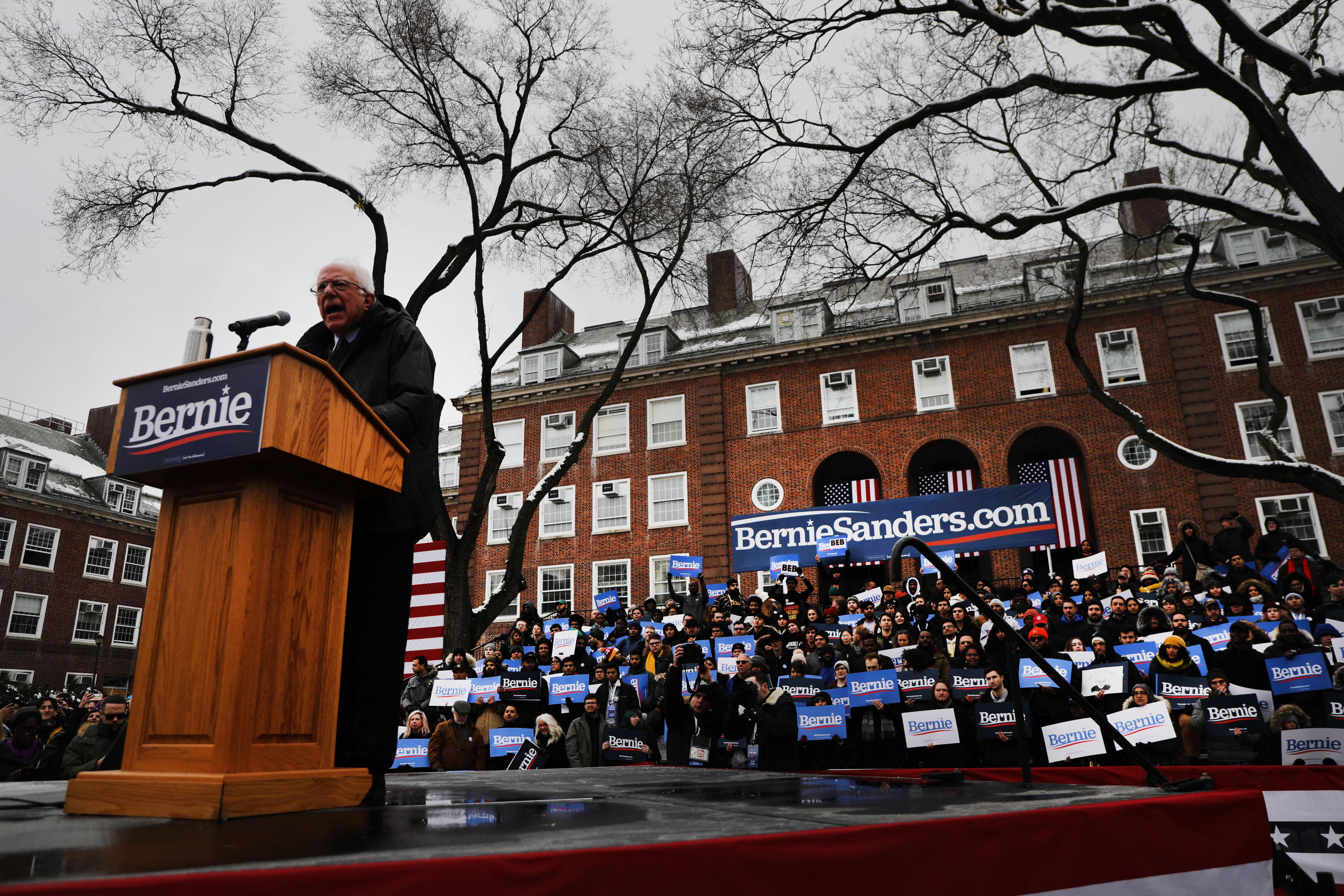 Bernie Sanders stands at a podium on a stage outdoors in front of supporters.