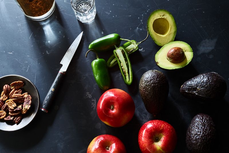 Three red apples, four avocados, and three jalapeño peppers lie on a black surface, along with a knife and a small bowl of pecans. One of the avocados and one of the peppers are each cut in half, lengthwise.