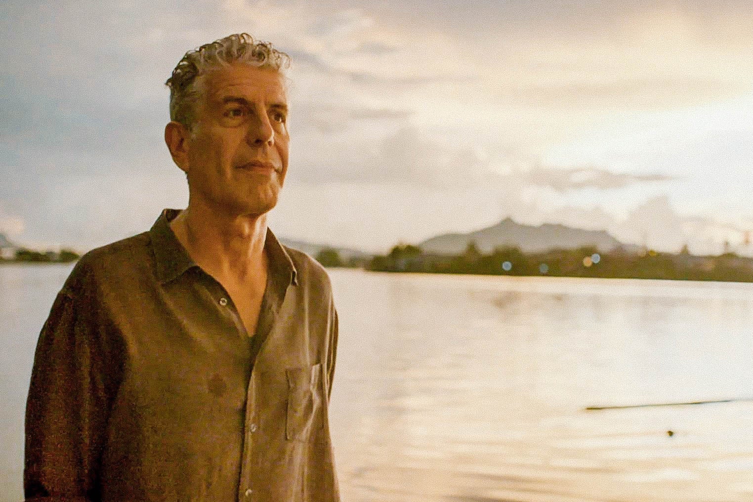 Anthony Bourdain stands in front of a body of water reflecting an orange sky.