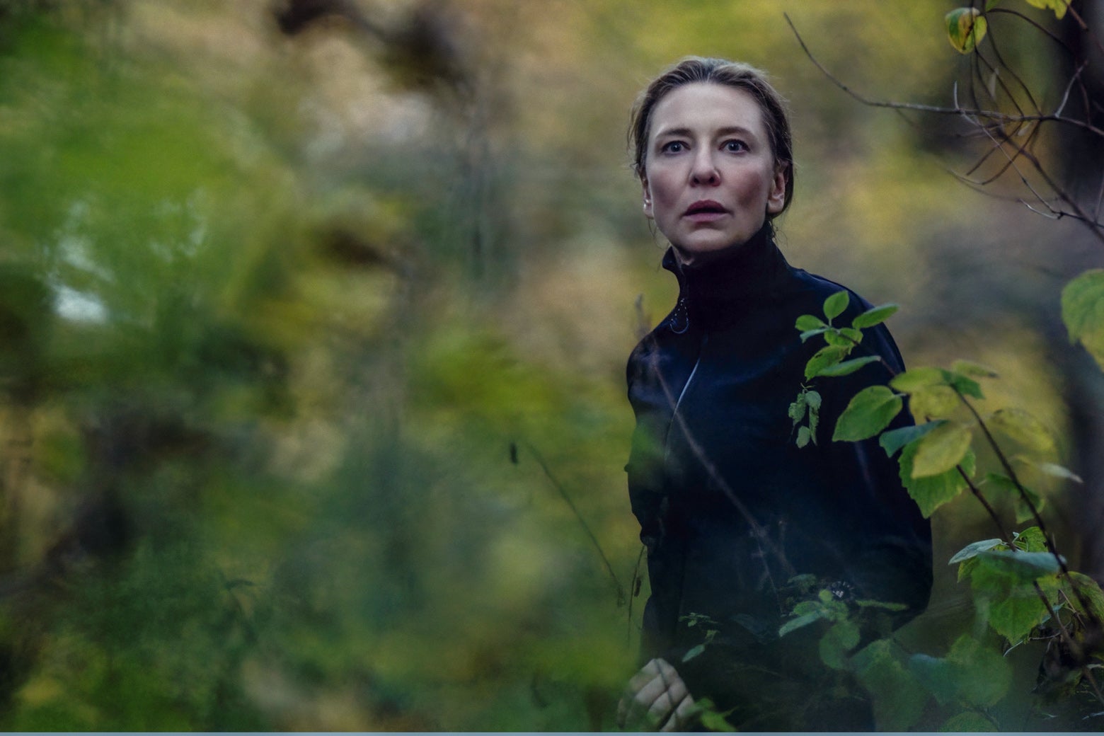 Lydia, wearing a black turtleneck, stares ahead in fear in the woods.