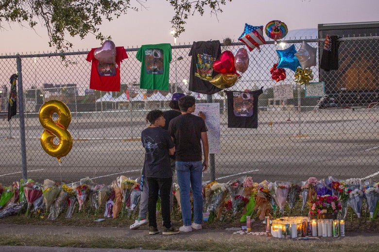 Two teens stand in front of a memorial of flowers, T-shirts, and balloons on a fence at dusk.
