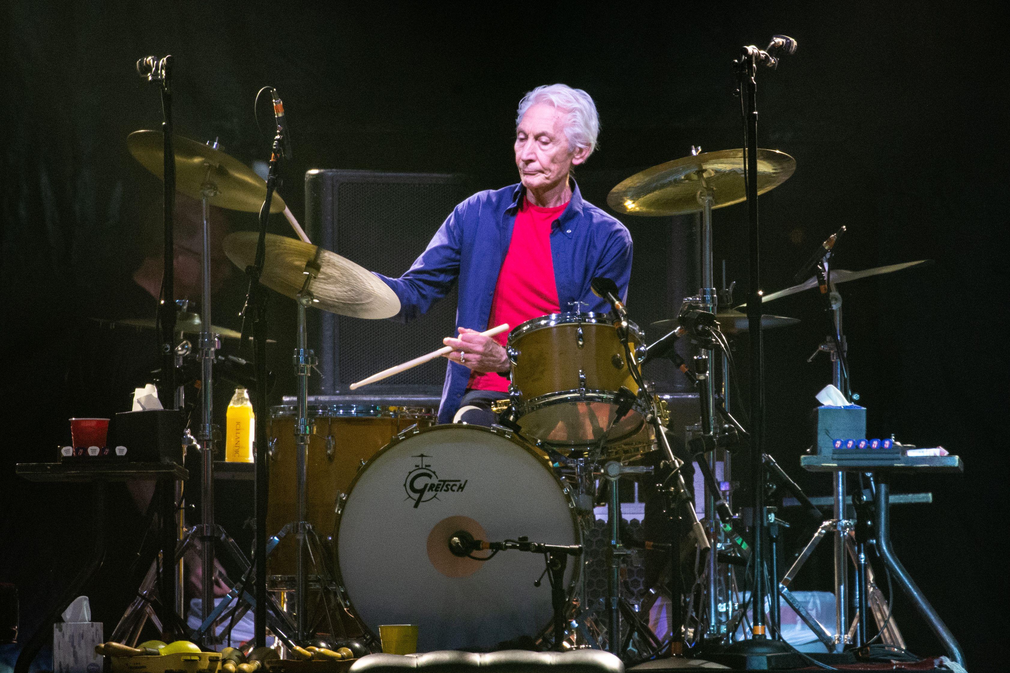 A man in a red t-shirt with a blue overshirt plays the drums on a stage. 
