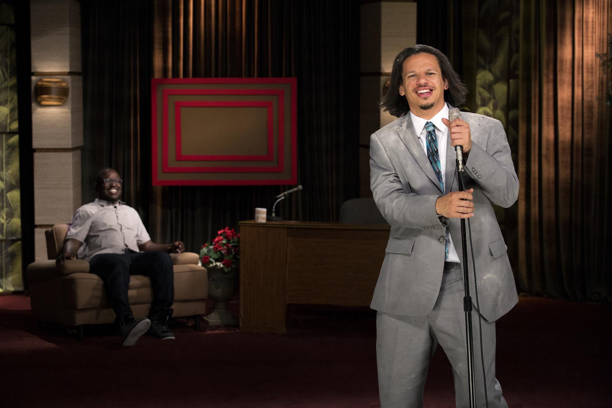 Eric Andre stands at a microphone with Hannibal Buress seated in an armchair in the background. 