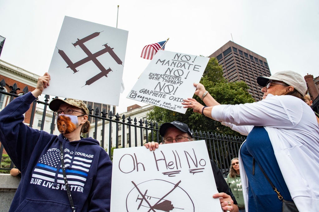 Three people outside the Massachusetts State House carry anti-vaccine signs, including one on which syringes form the shape of a swastika.