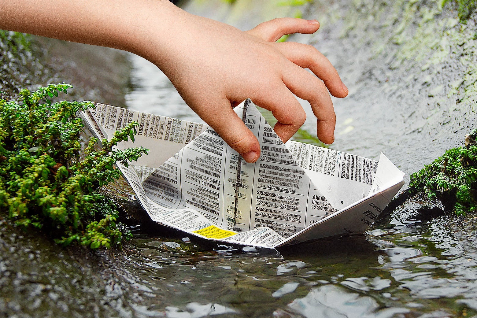 A child's hand gently pulling a newspaper boat out of a gutter.