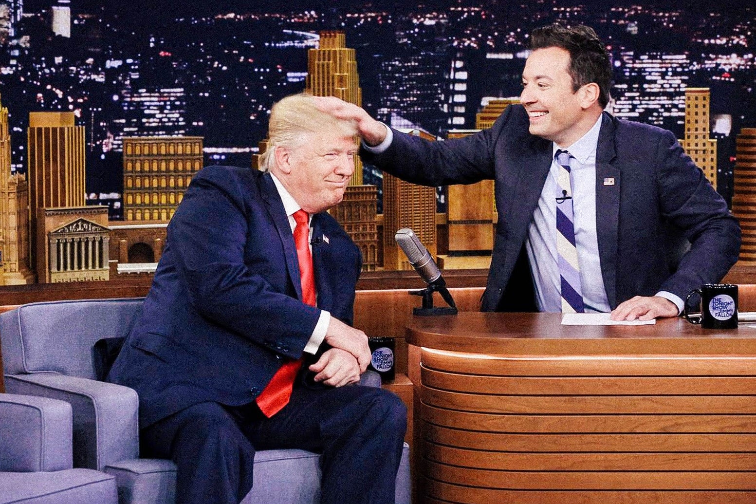 A smiling Jimmy Fallon musses Trump's hair with his hand.