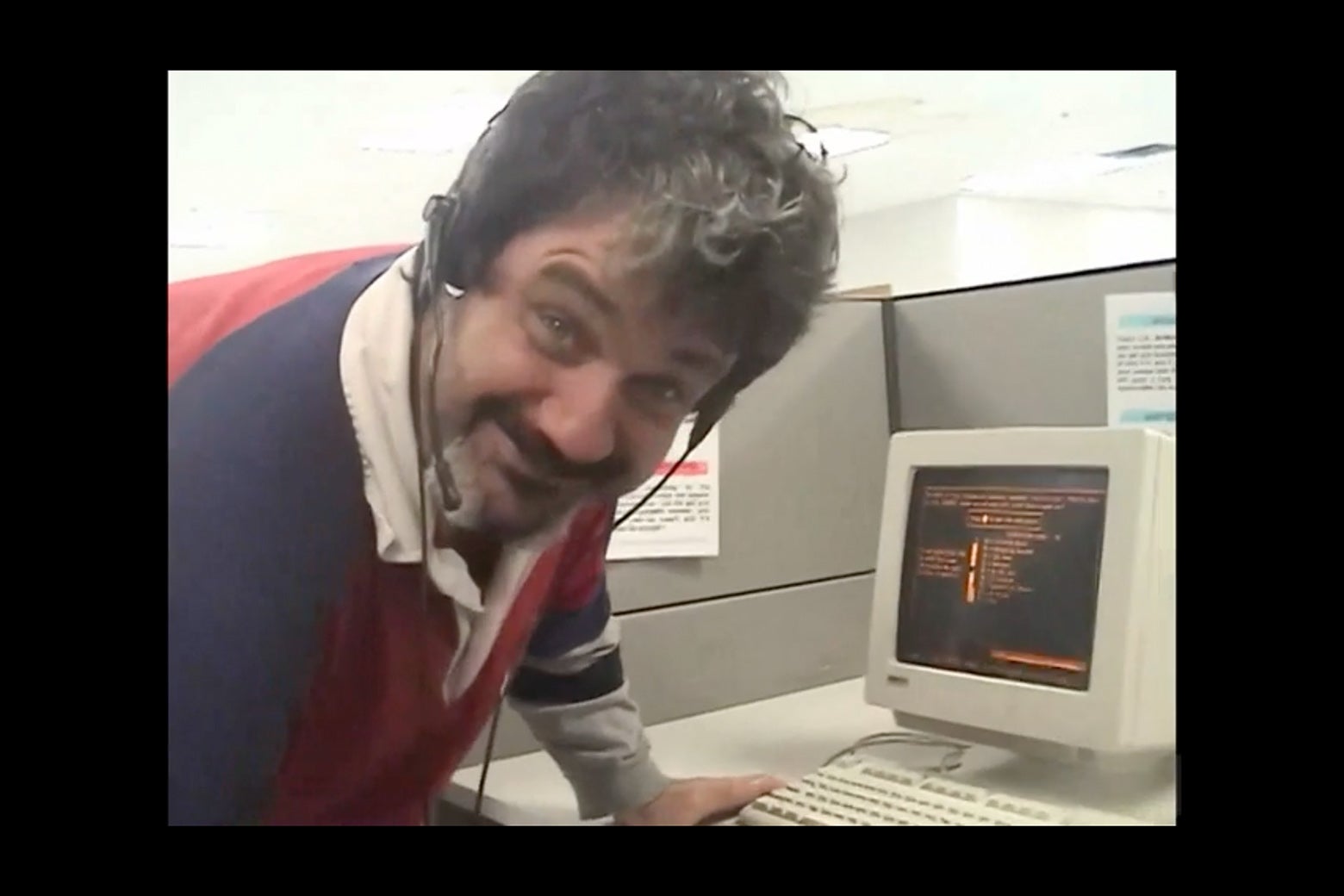 A man in a rumpled polo shirt, graying curly hair, and a beard, stoops in front of a very 1990s-looking computer, wearing a headset, making a sort of smirking expression into the camera as if to say, "Yeah, I know it's not great."
