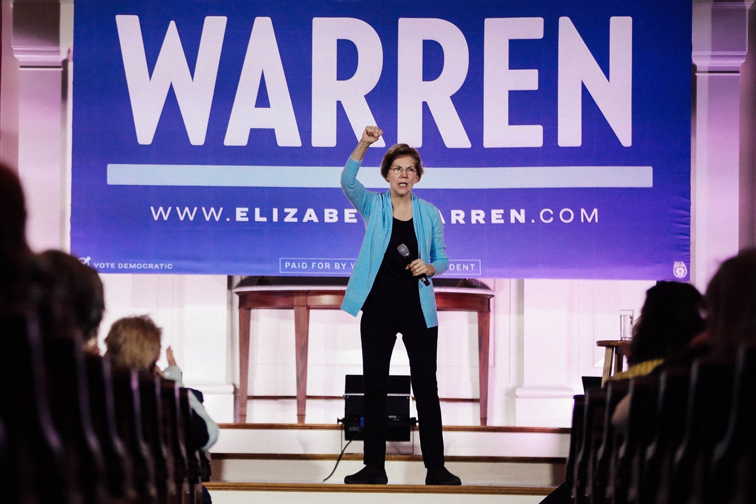 Elizabeth Warren raises her fist and holds a mic in front of a banner bearing her name at a campaign event.