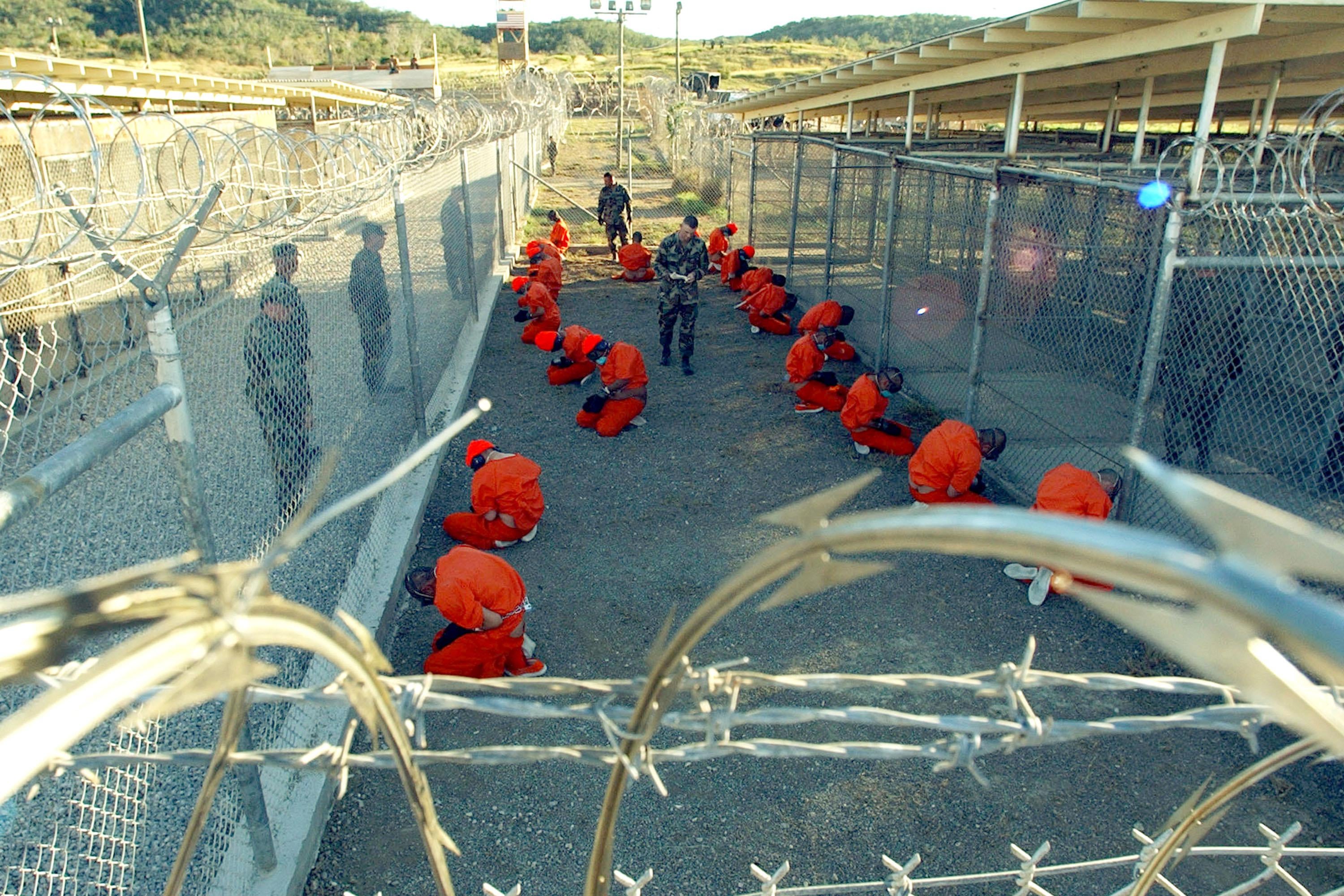 Men in orange jumpsuits kneel in an enclosed area.