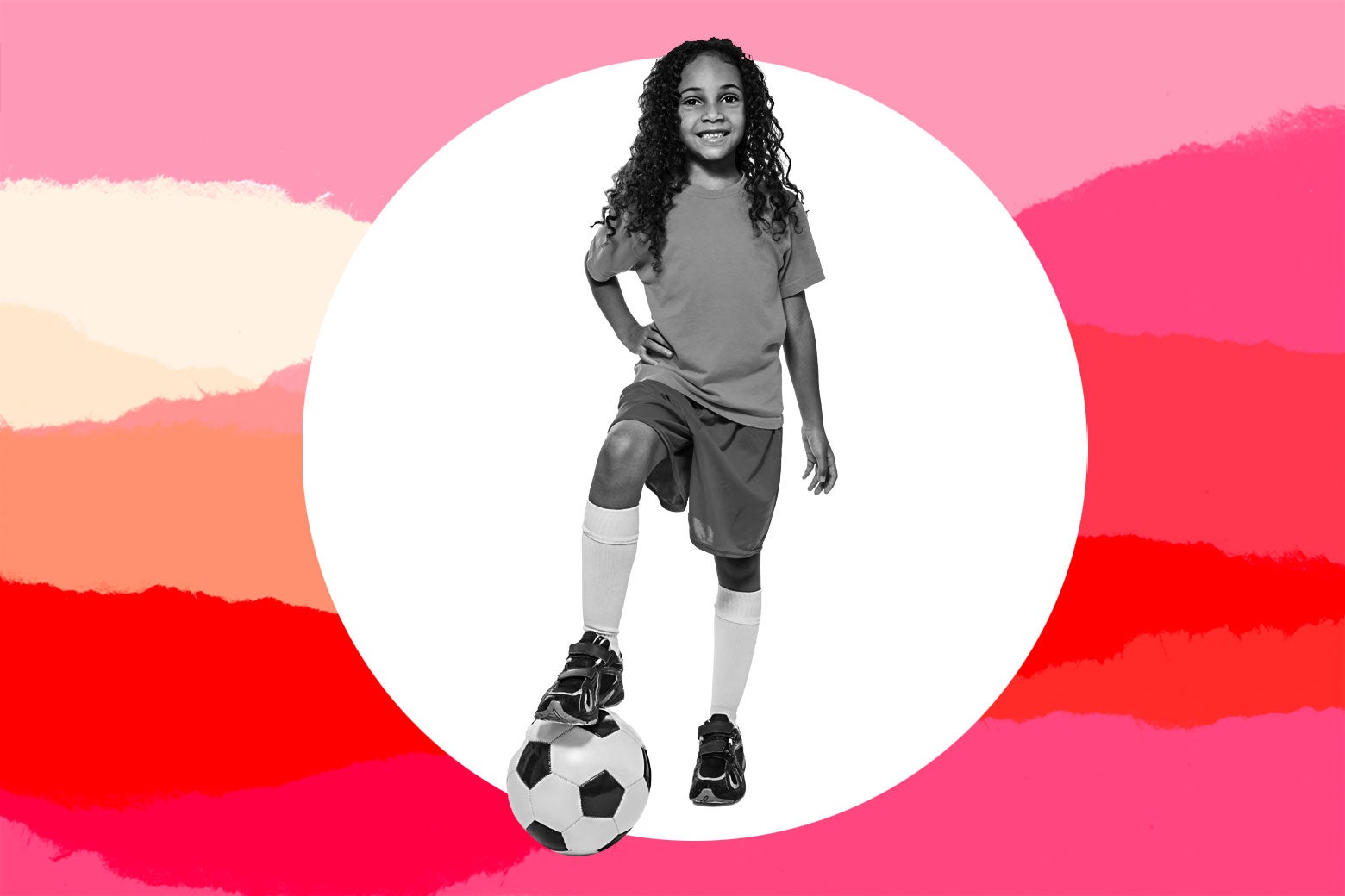 A girl in soccer gear smiles as she puts her foot on a soccer ball.