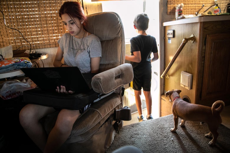 A teenage girl works on a laptop, sitting in an easy chair inside a cluttered mobile home. Behind her a dog looks out the open door in which a boy is standing.
