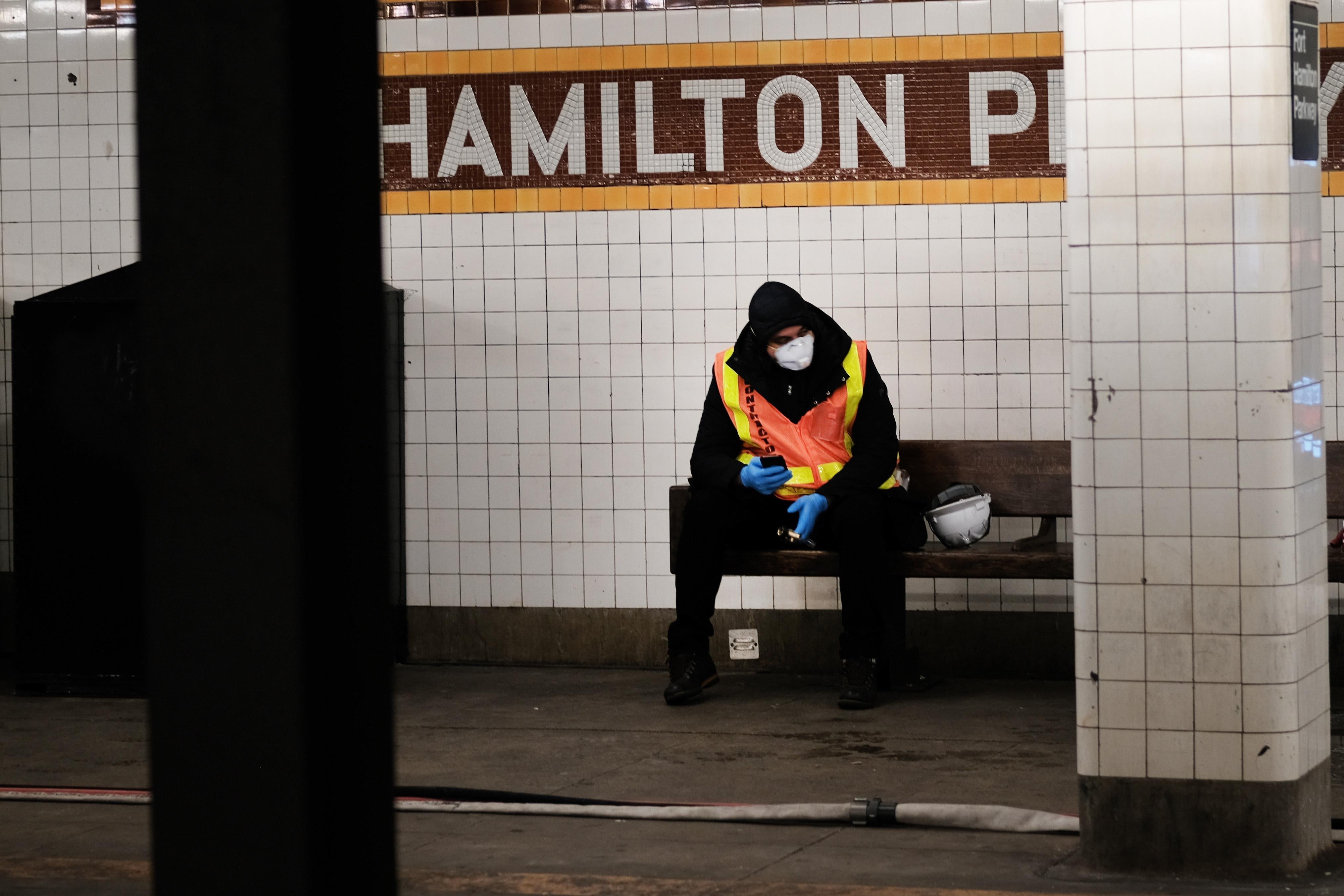 A worker wearing an orange vest and a face mask sits on a bench at a subway station.