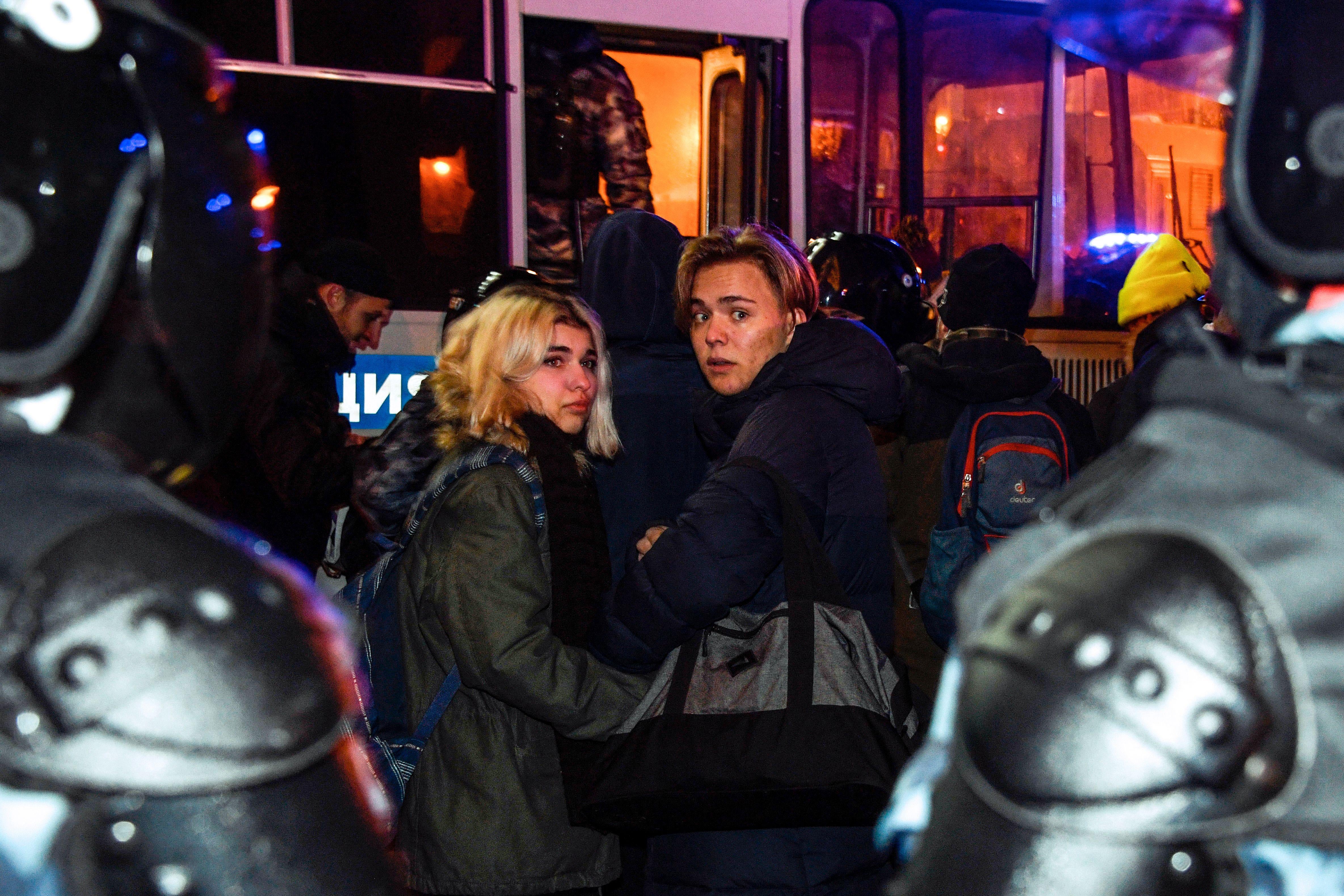 Two women flanked by armored police officers look back toward the camera as they are forced onto a police bus. 