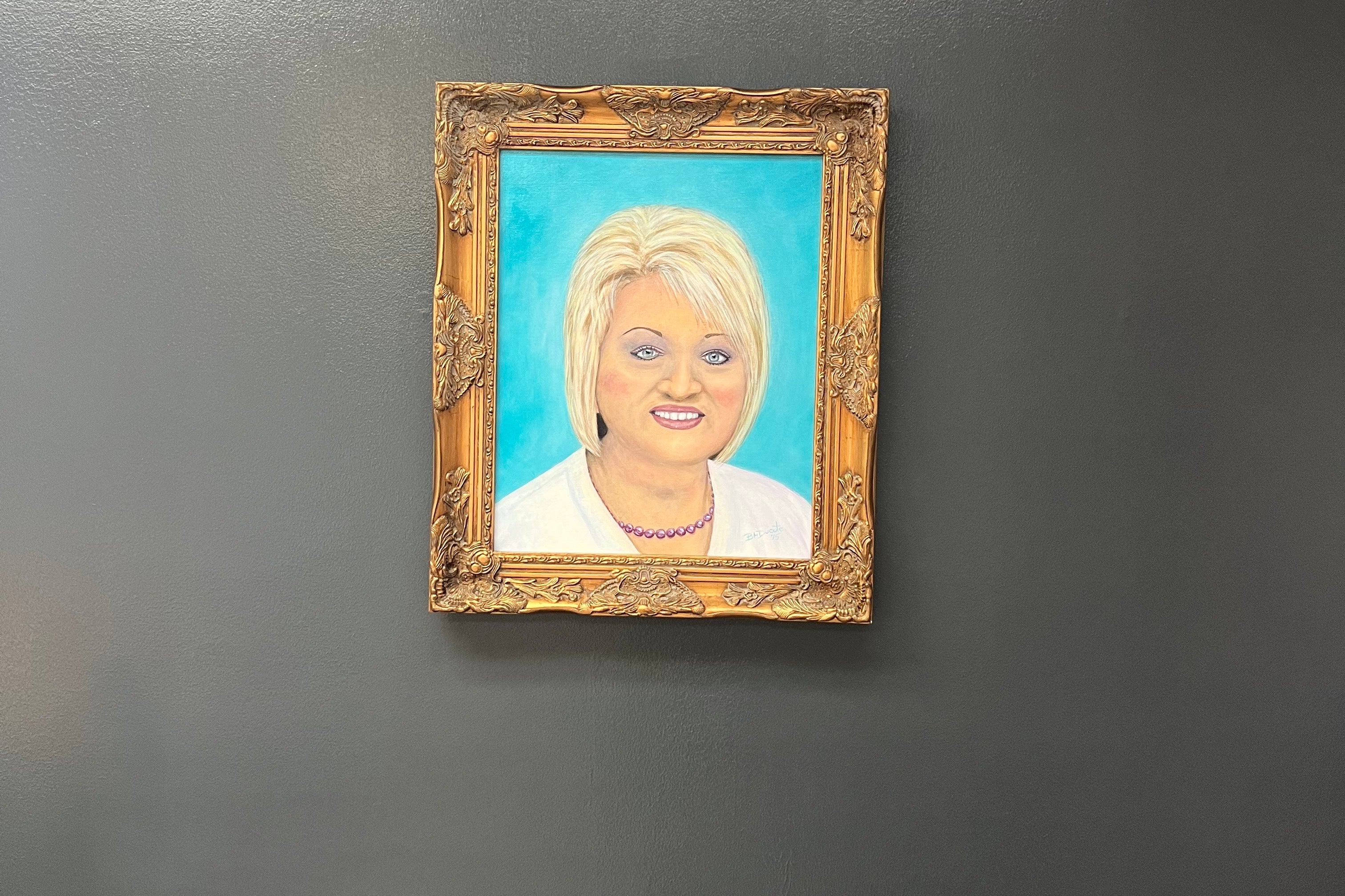 A woman with a blond bob smiles in a gold-framed portrait, with a teal background.