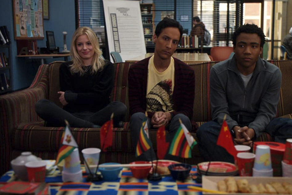 Gillian Jacobs, Danny Pudi, and Donald Glover sit on a couch, staring a table full of small Chinese and Ghanian flags.