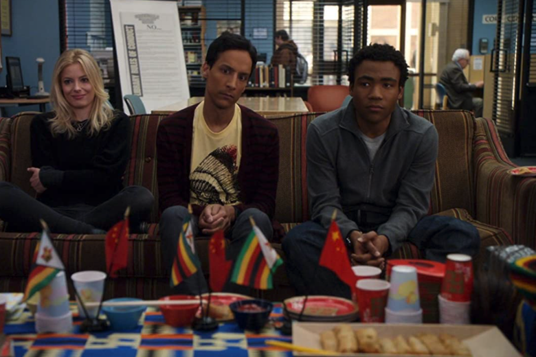 Gillian Jacobs, Danny Pudi, and Donald Glover sit on a couch, staring a table full of small Chinese and Ghanian flags.