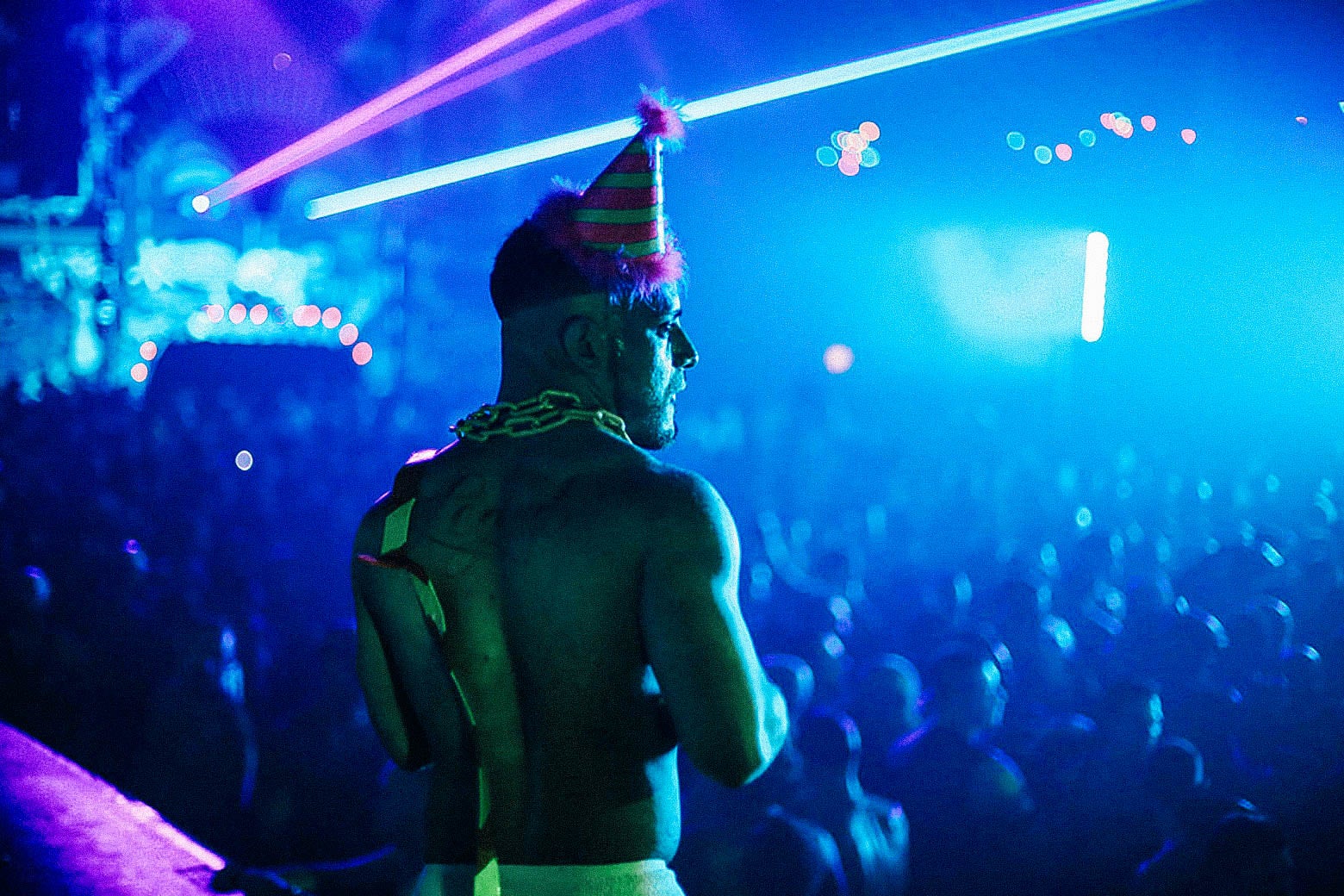 A topless man wearing a party hat and a chain around his neck stands above a crowd of revelers on the dance floor. Laser beams punctuate the dark scene.