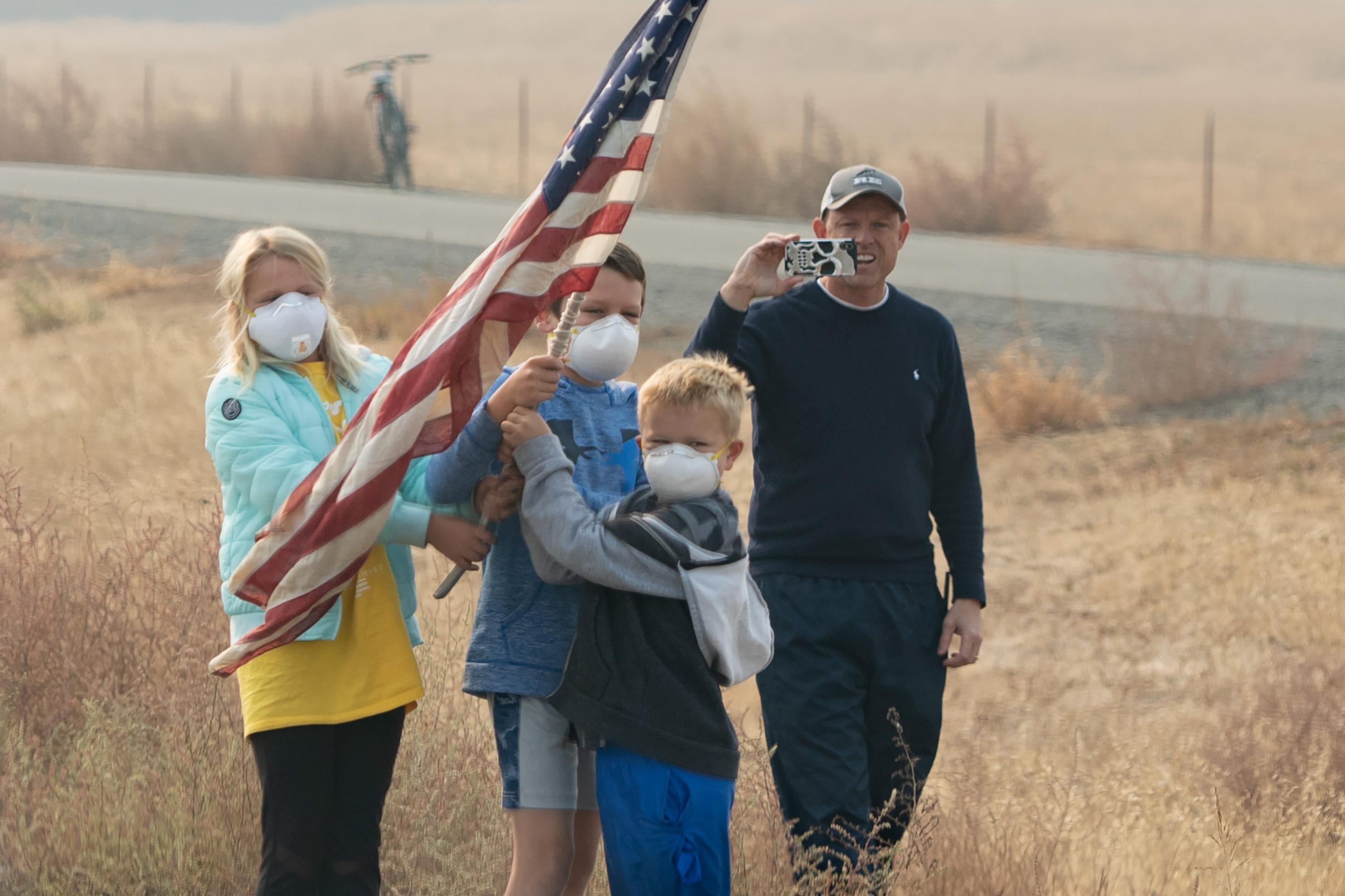 Three children wearing white N95 masks hold up an American flag while a man stands behind them smiling and holding up a phone to take a picture or video.