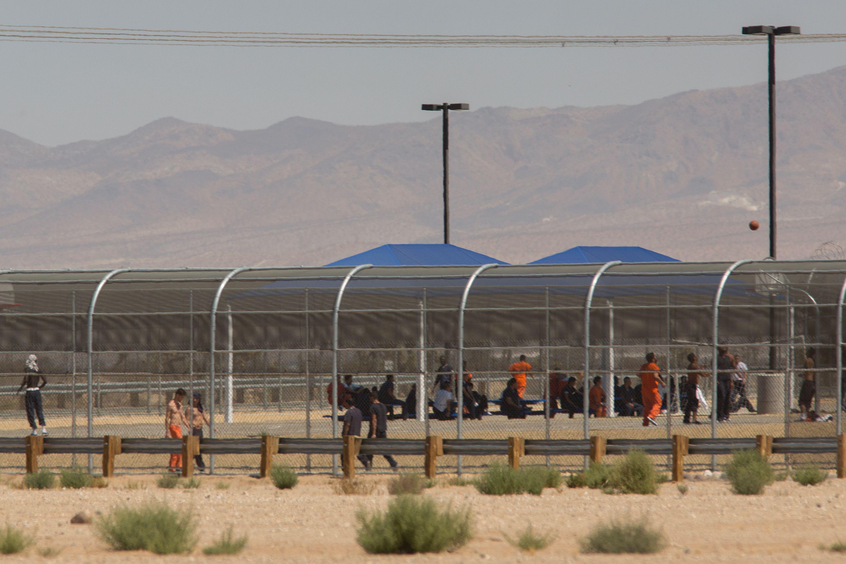 Adults in prisonlike clothes can be seen behind a fenced-off facility in the desert with mountains in the background.