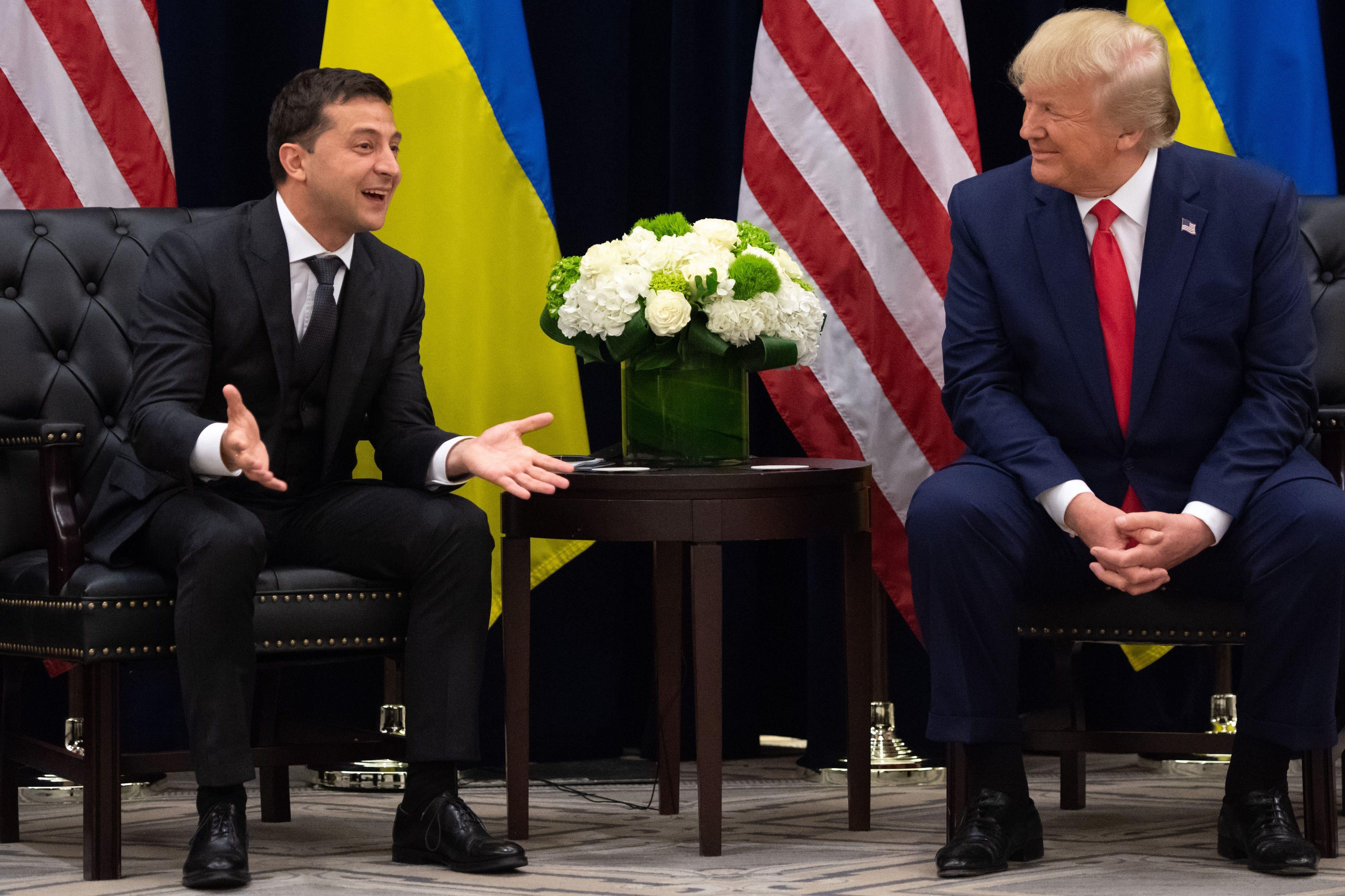 Zelensky smiles and gestures beside Trump smiling at him, both men seated with Ukrainian and U.S. flags behind them.