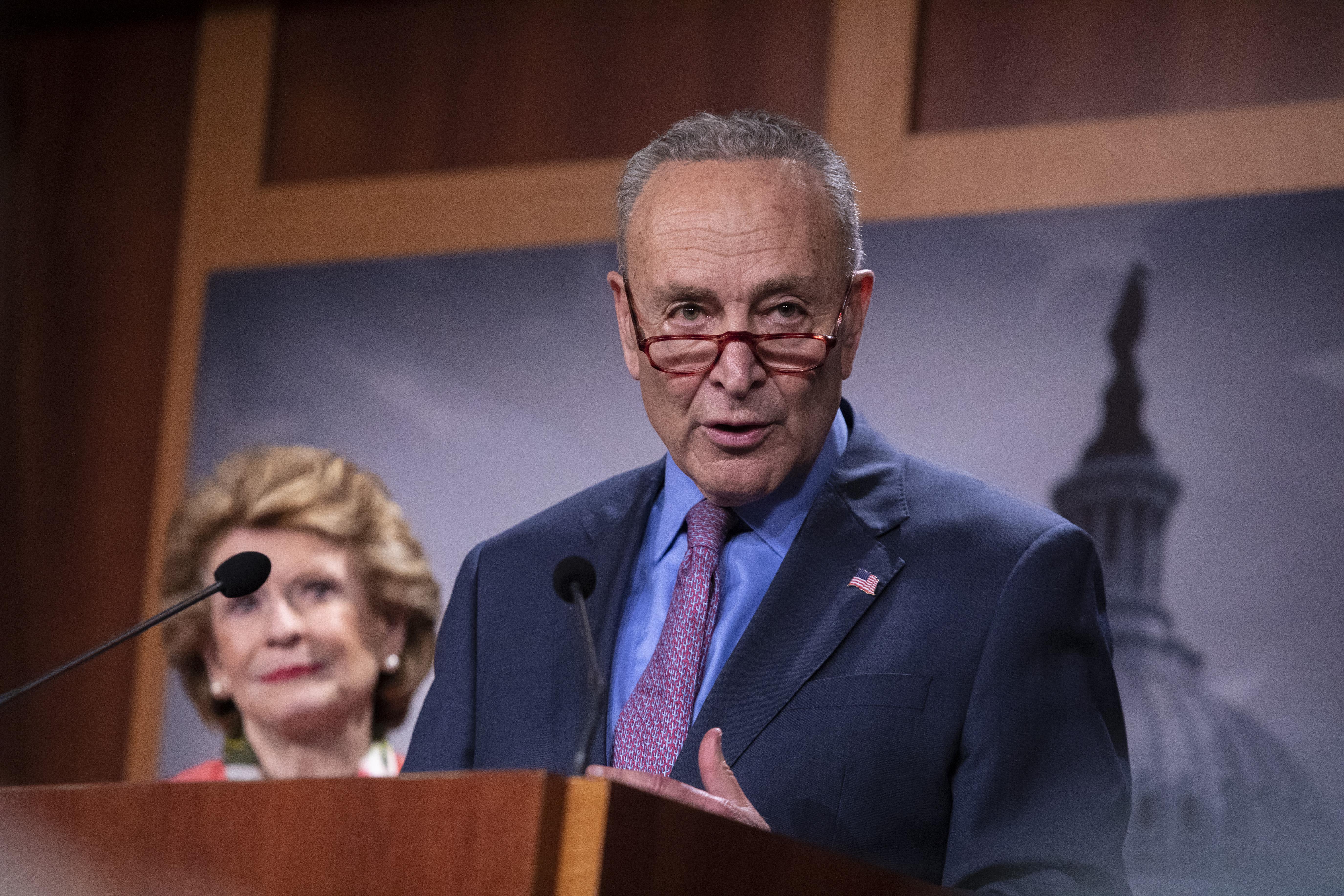 Chuck Schumer speaks at a lectern as Debbie Stabenow stands behind him.