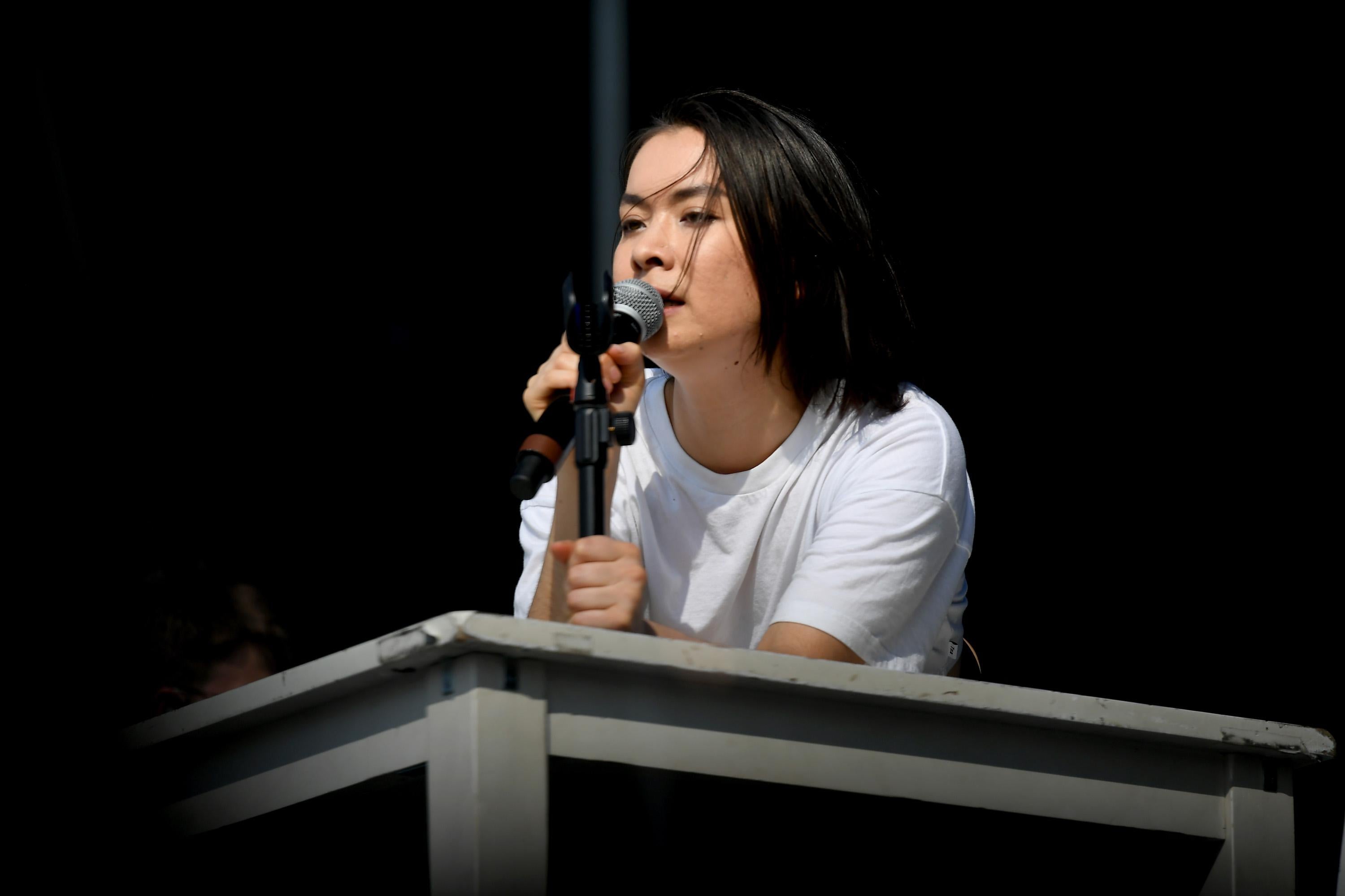 A woman with short black hair wearing a white T-shirt sings into a microphone in her hand. She is leaning on a gray table onstage.