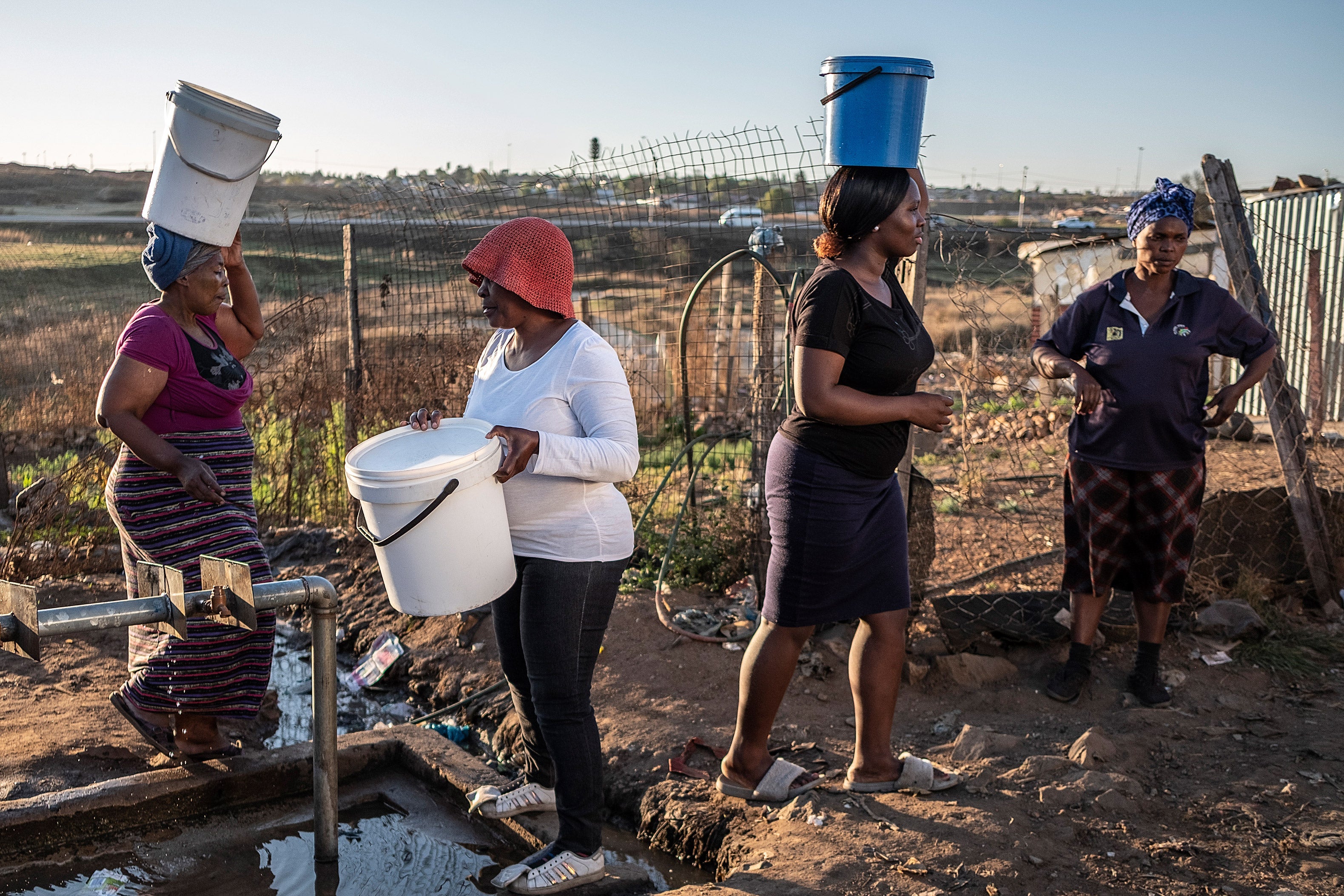 Women with buckets stand in line to get water from a tap.