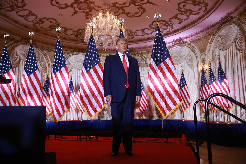 Trump, wearing a blue suit and red tie, stands under a ballroom chandelier on a red-carpeted stage lined with American flags.