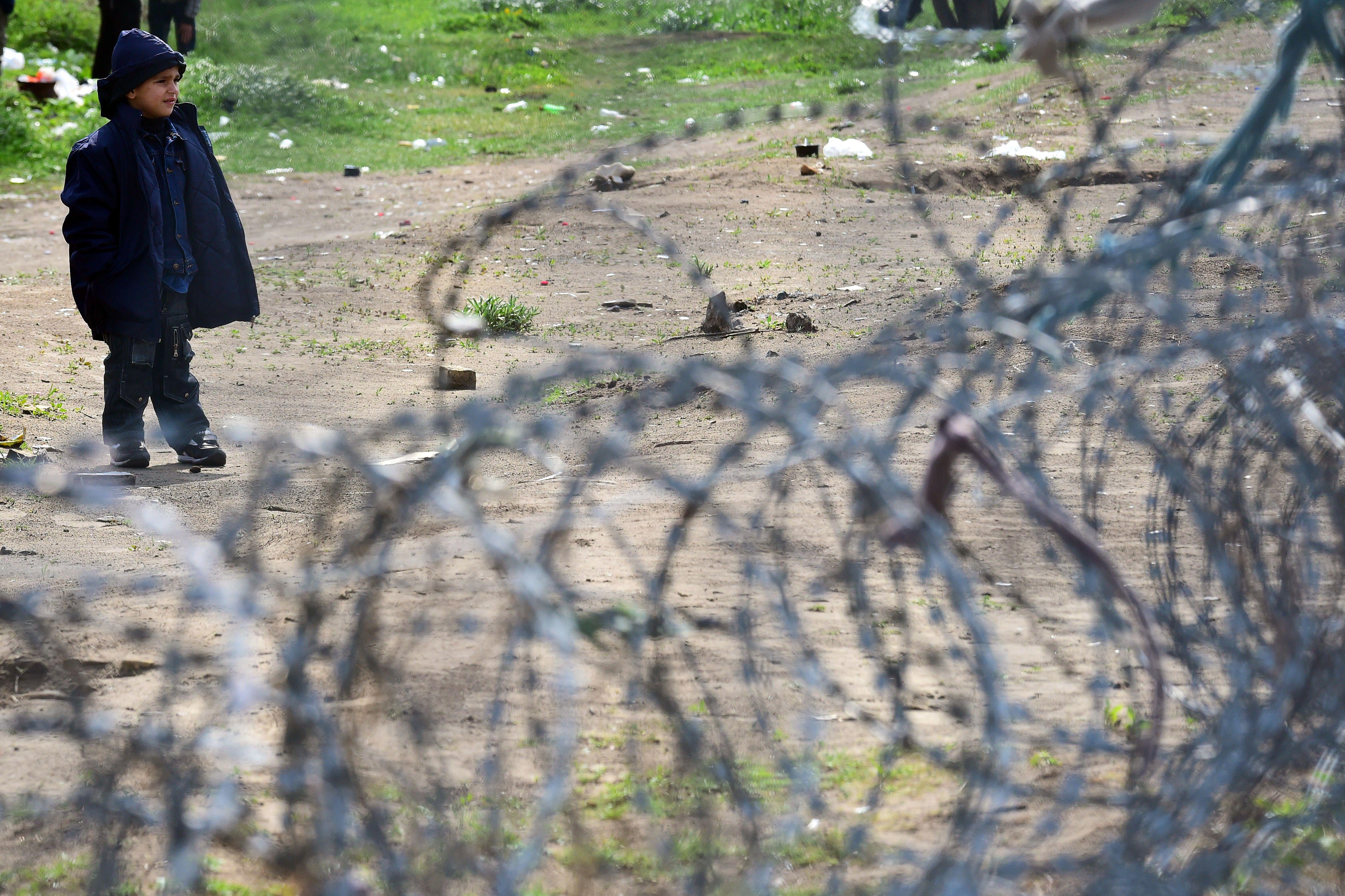 A young migrant boy stands behind a barbed wire fence.