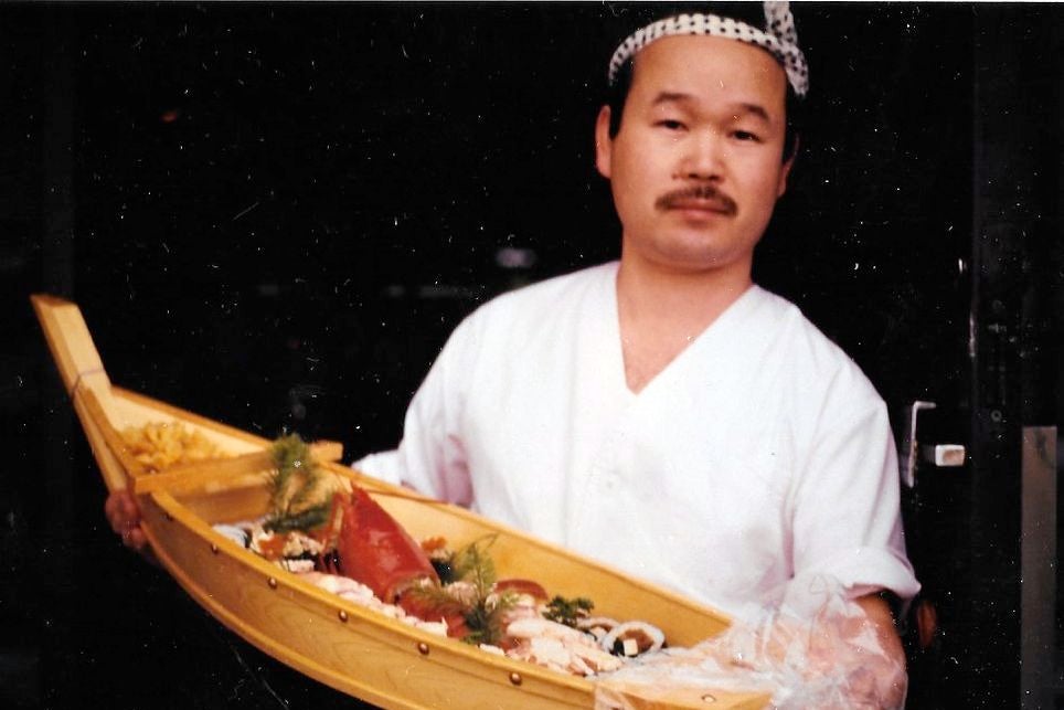 A mustached man holds a long, boat-shaped bowl filled with sushi.
