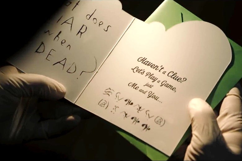Rubber-gloved hands hold open a greeting card. The left side has "What does a liar do when he's dead?" written on it, while the left side has "Haven't a clue? Let's play a game, just me and you" printed above a ciphertext made from random symbols.