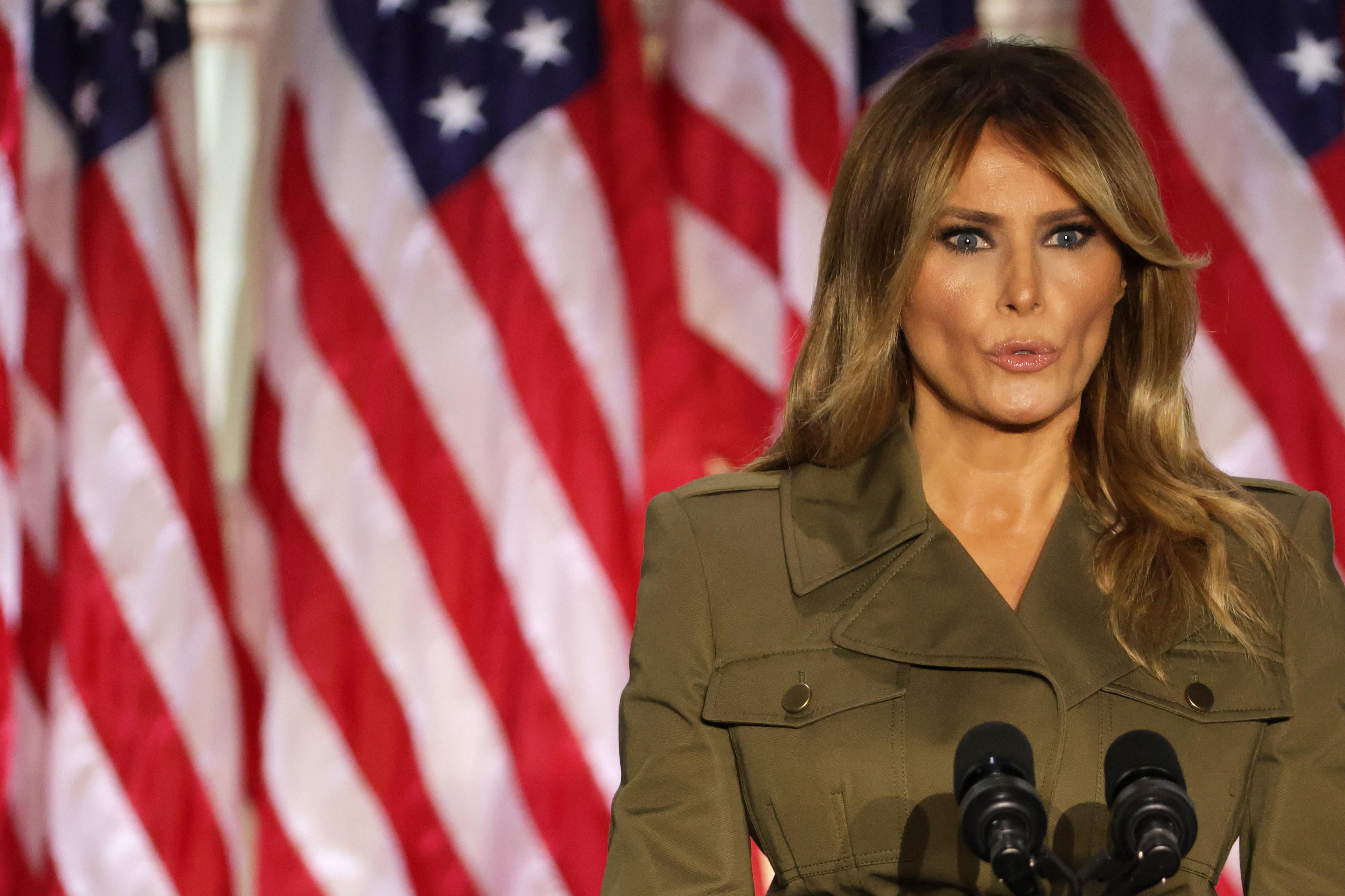 Melania Trump, in an olive-green military-looking jacket, speaks into a cluster of microphones, backed by several U.S. flags.