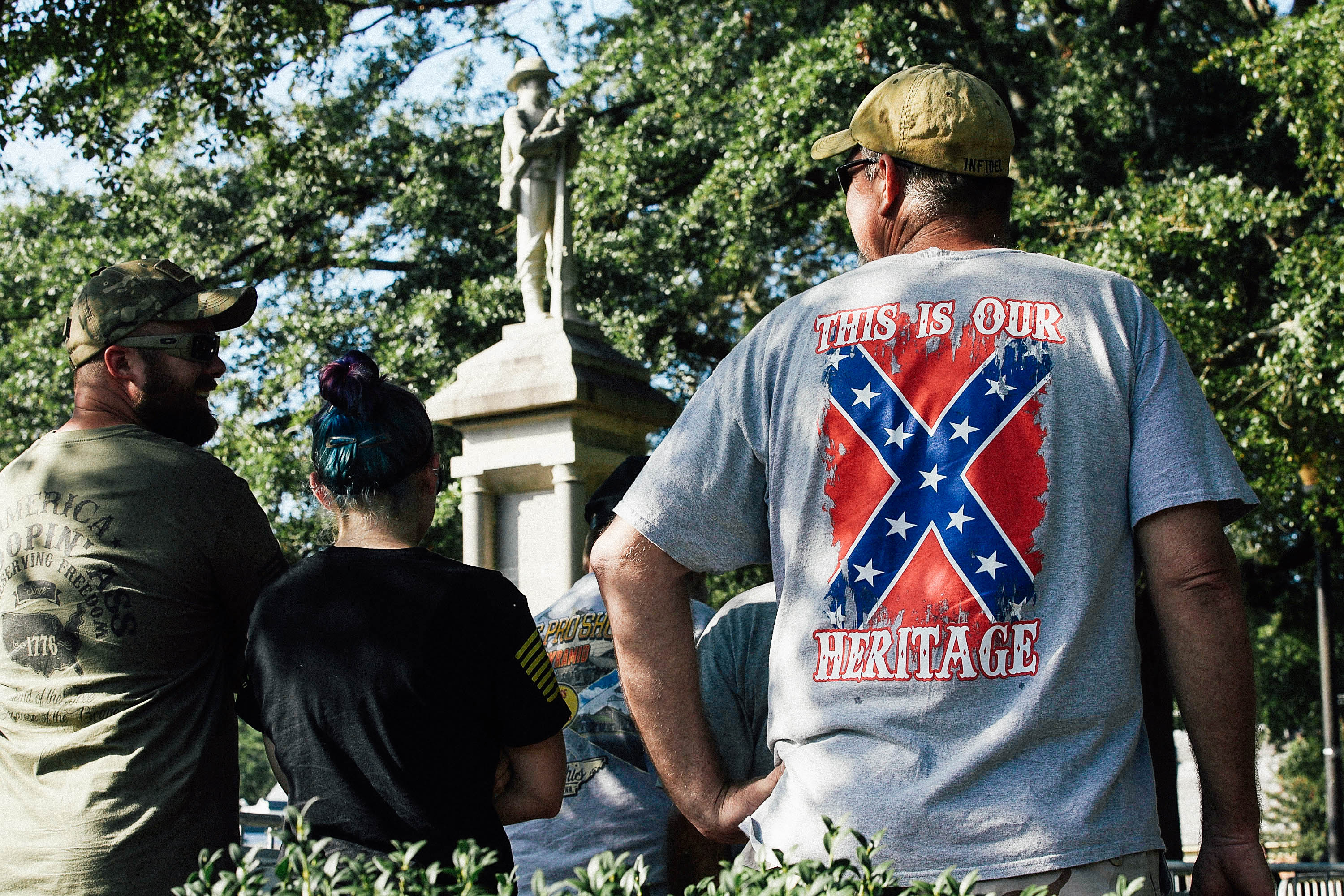 A man wearing a T-shirt with the words "This is our heritage" around the Confederate battle flag stands in front of a Confederate statue among other statue supporters.