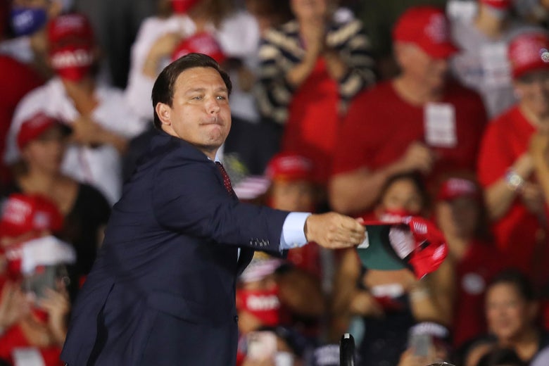 Ron DeSantis, wearing a suit as he stands in front of a MAGA crowd, tosses a red hat.