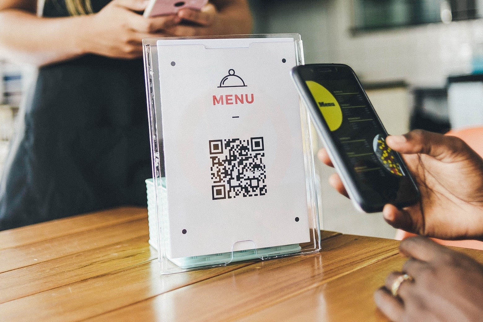 A QR code on a sheet of paper labeled "MENU" standing upright on a table. Someone holds their phone up to the code to scan it, displaying the menu on the smartphone screen.