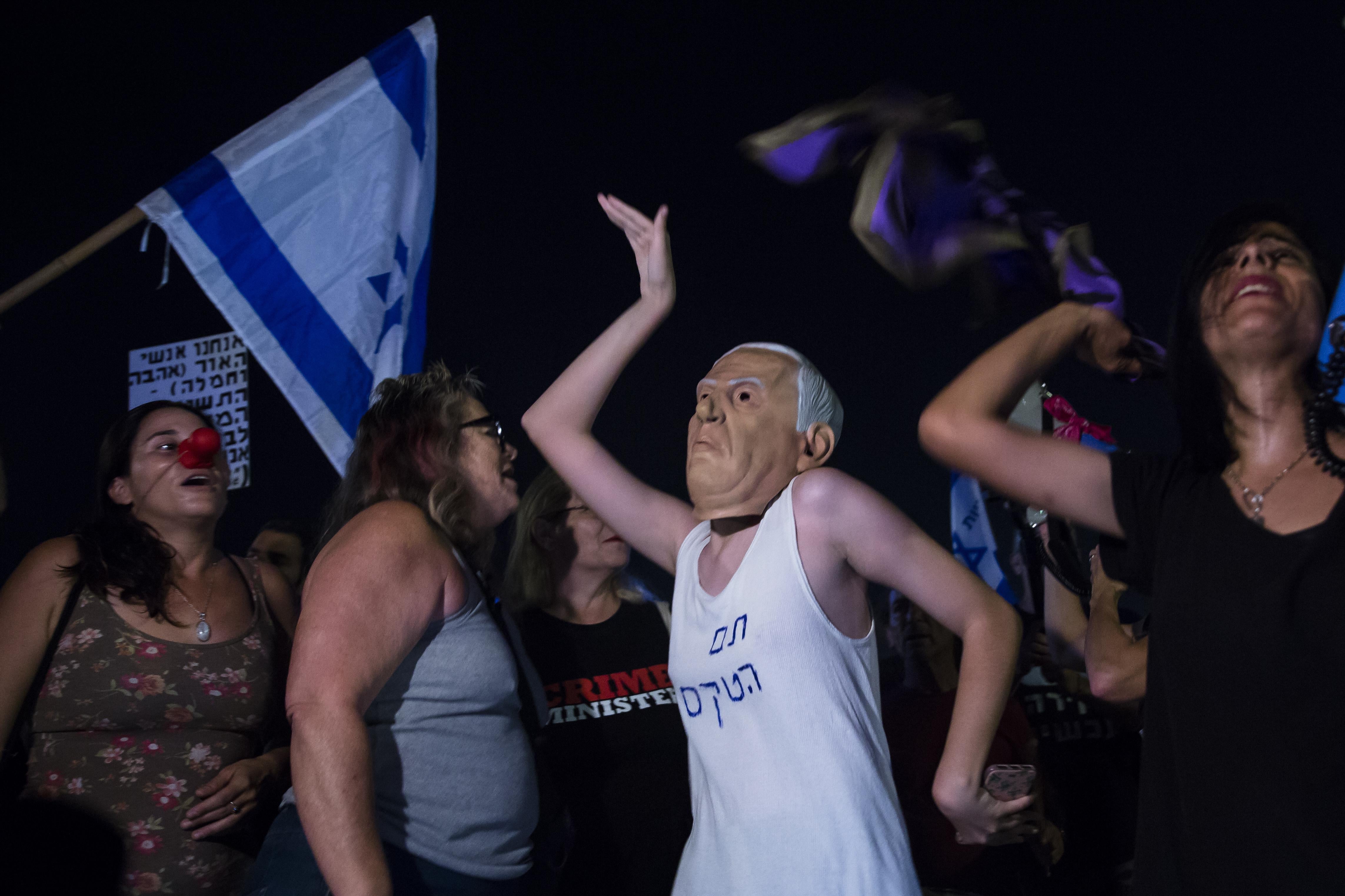 A young woman, wearing a rubber mask of Benjamin Netanyahu over her head, dances in a crowd of people.