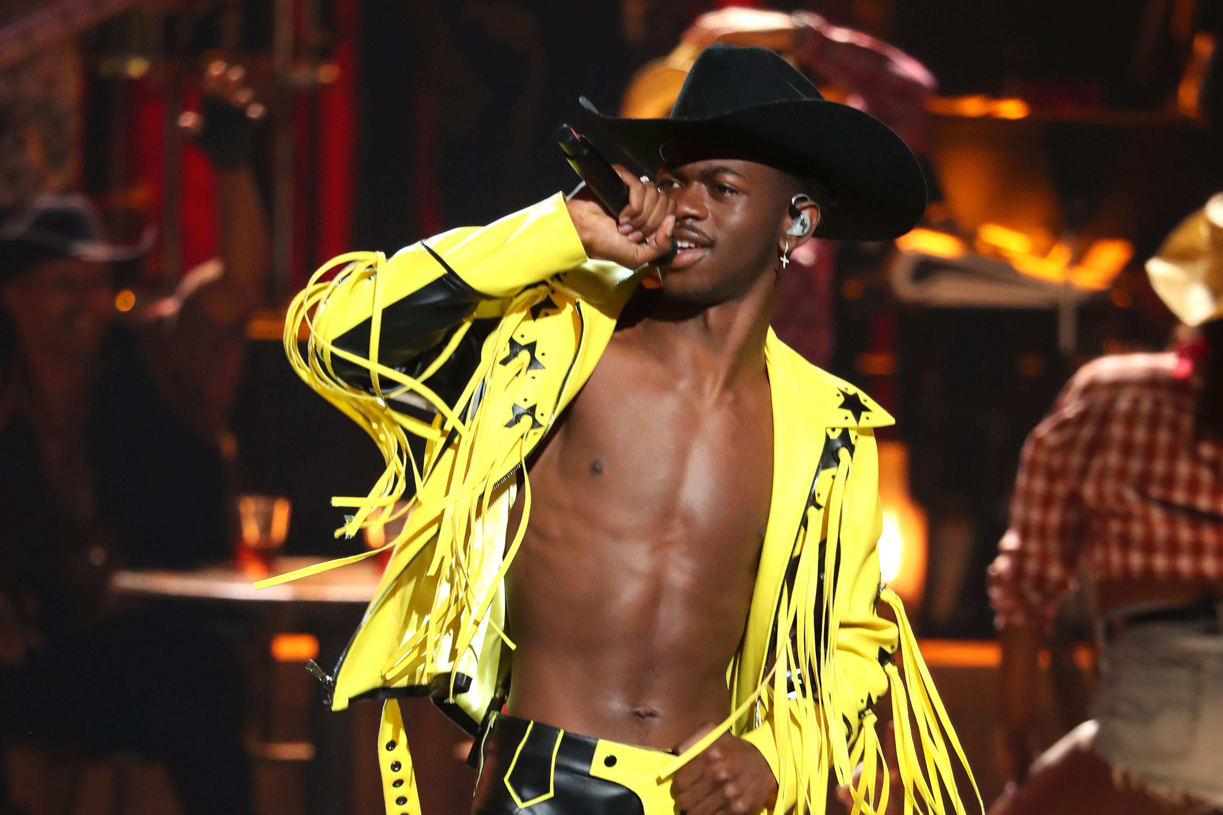 Lil Nas X in a yellow leather fringed jacket, a black cowboy hat, and no shirt.